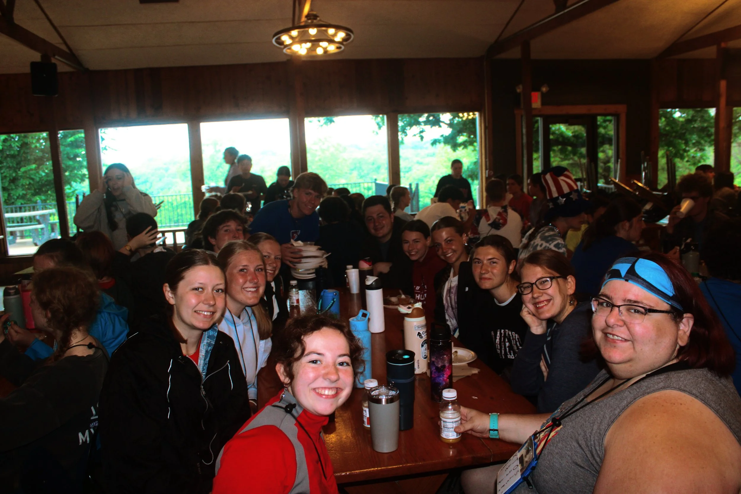 A group of smiling young people at a table indoors, with some holding water bottles and others eating, in a cozy wooden cabin with large windows showing trees outside.
