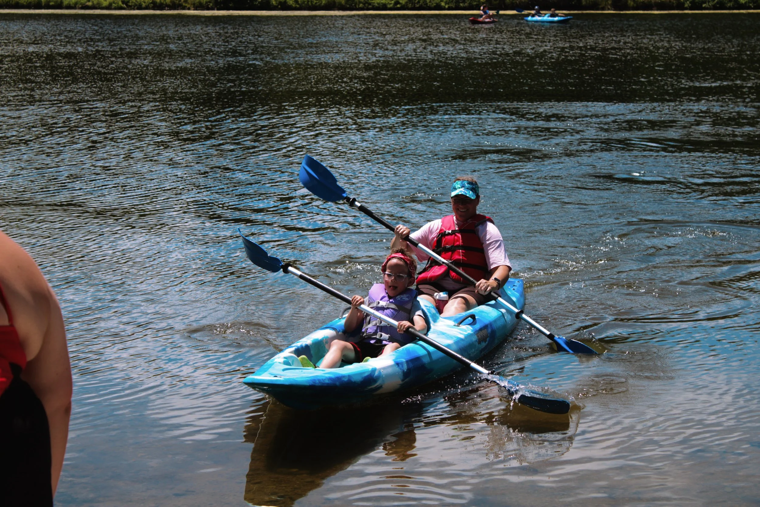 A woman and a young girl sit in a blue and white tandem kayak on a body of water, each holding a paddle with black shafts and blue blades, enjoying a sunny day. In the background, other kayakers are visible on the water, and the shoreline with trees 