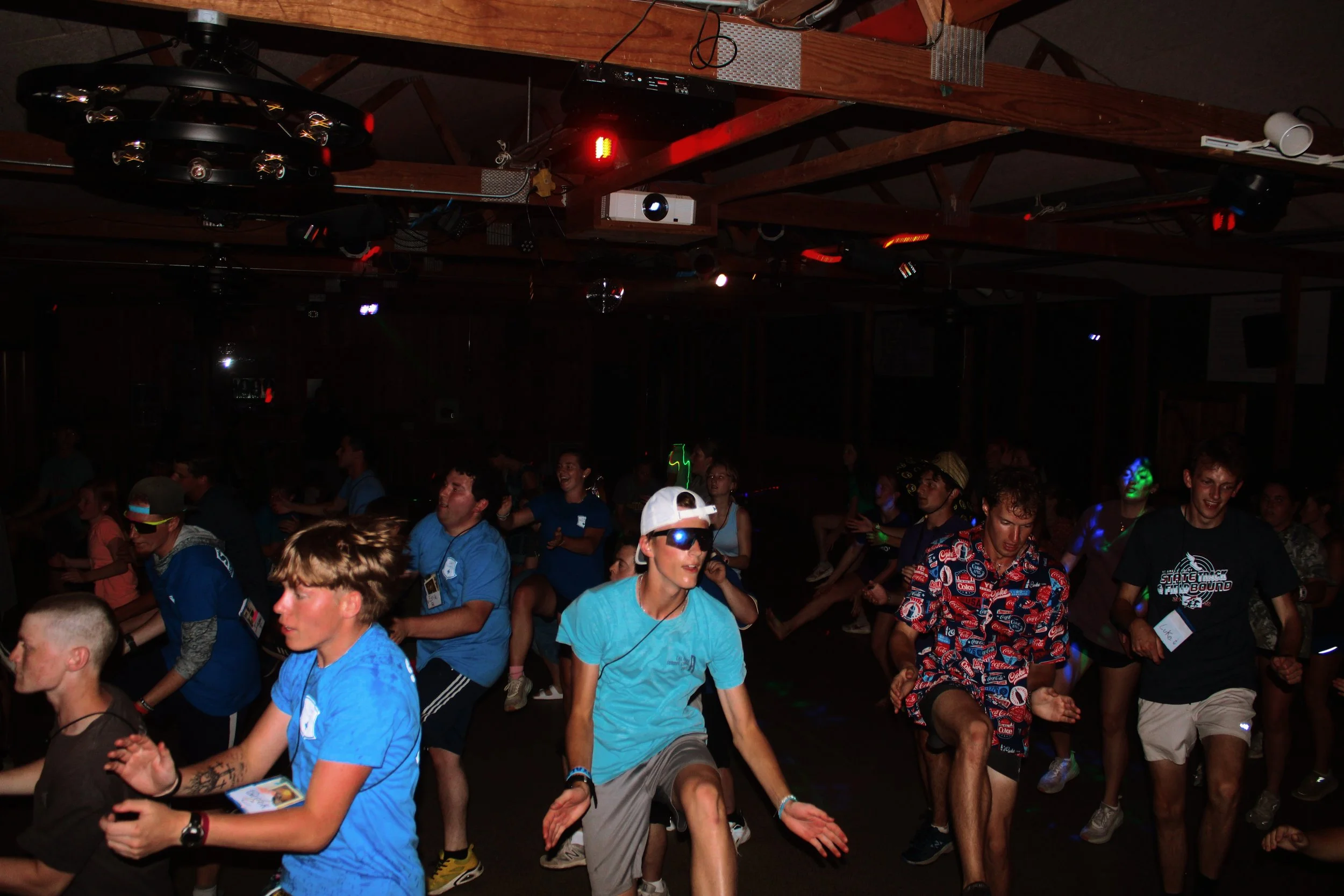 A group of young people dancing indoors in a dark room, with some wearing bright clothing and sunglasses, under dim colored lighting.