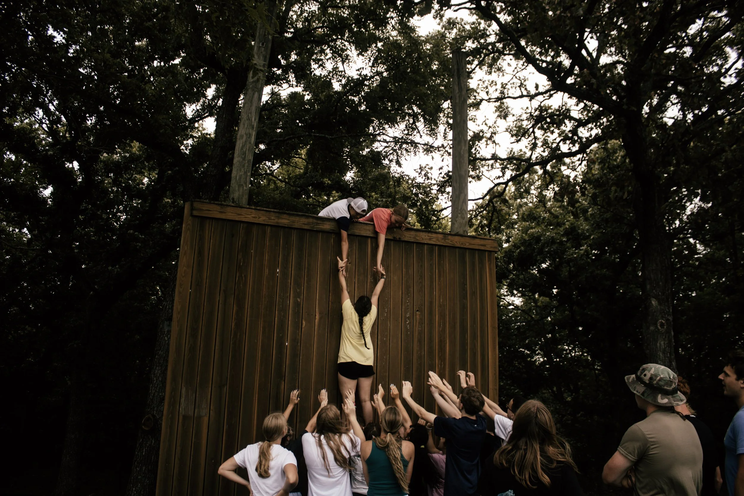 A group of children cheering as two kids high-five on top of a wooden obstacle during an outdoor event in a wooded area.