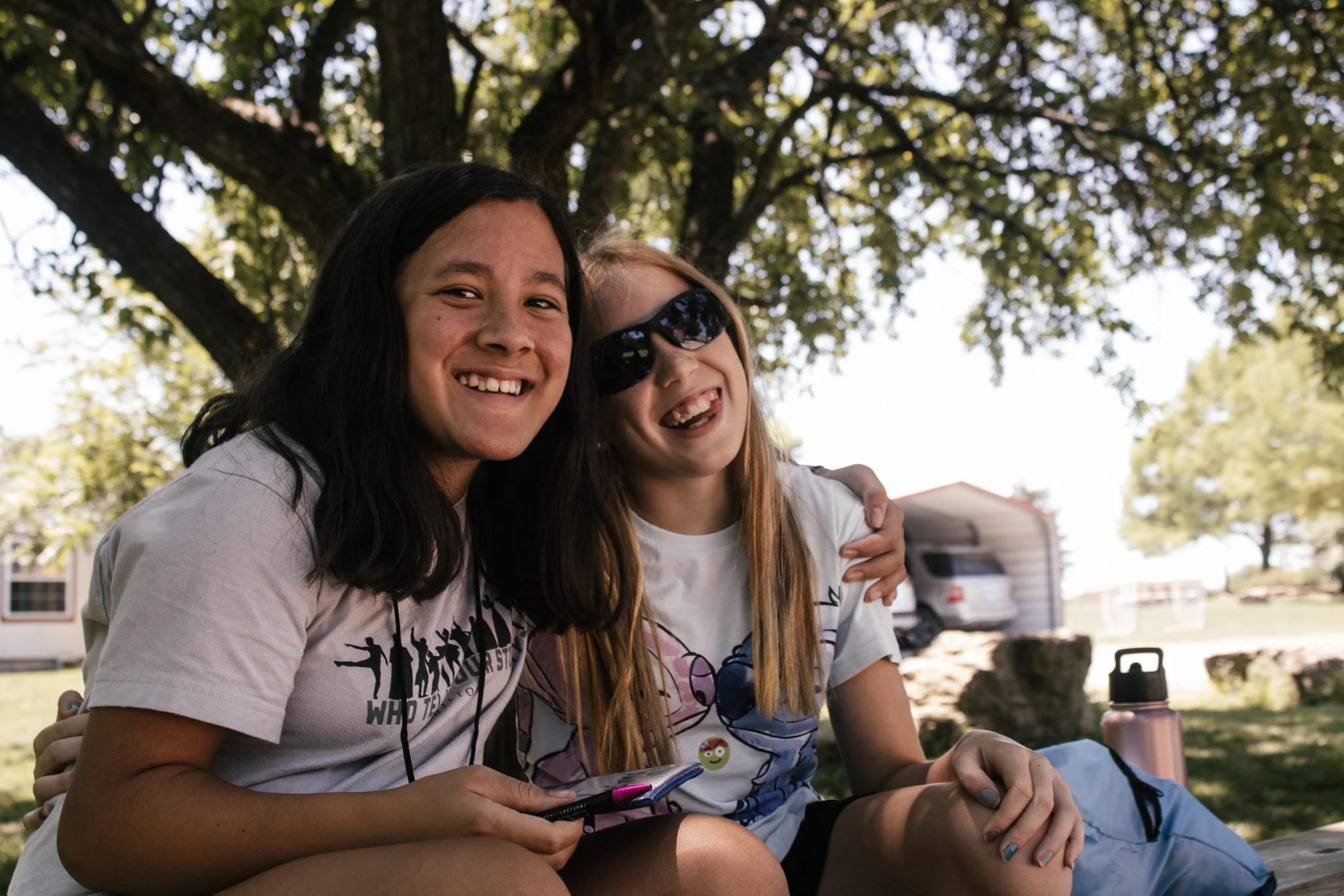 Two young girls sitting outdoors under a tree, smiling and hugging each other.