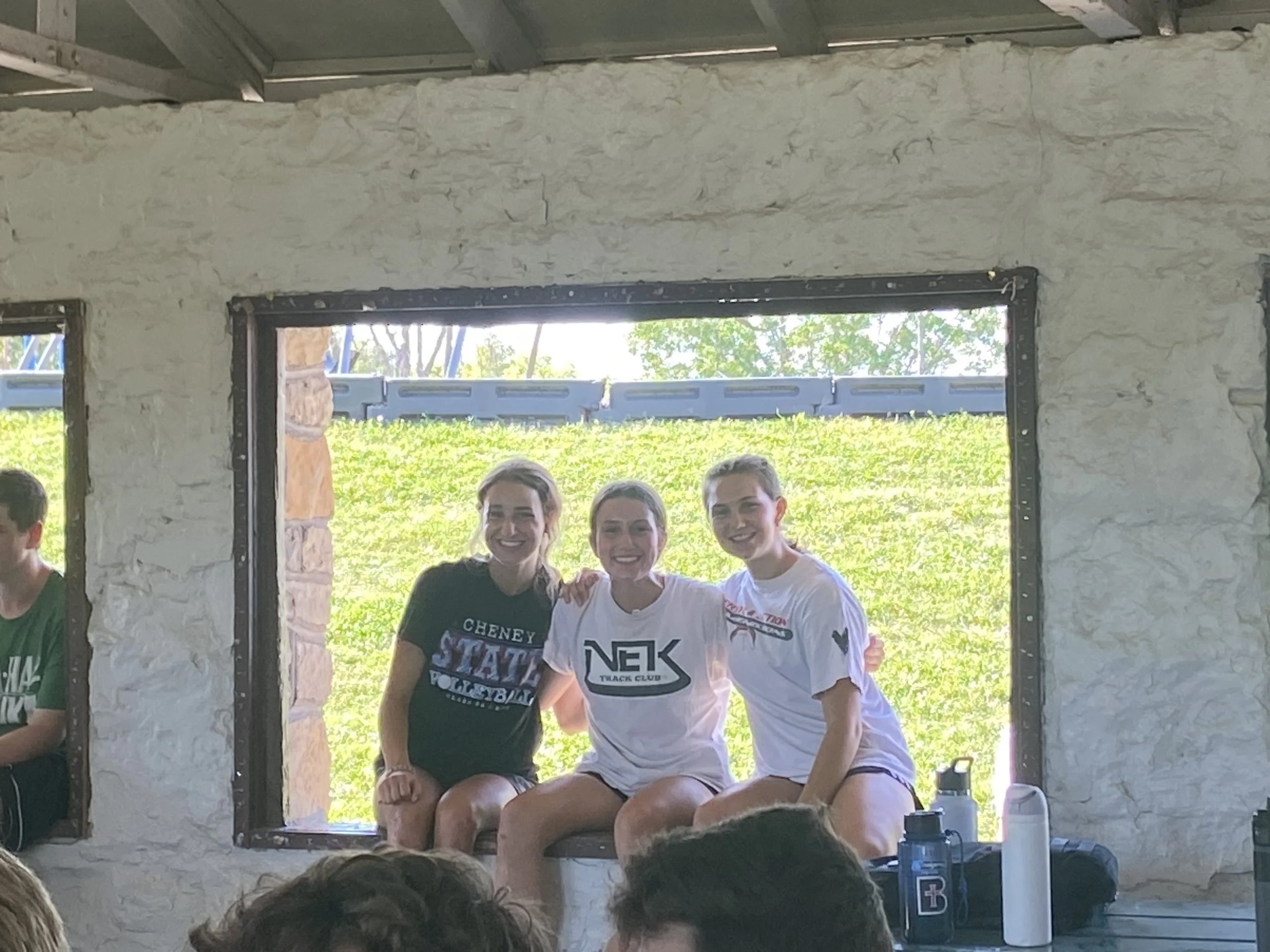 Group of four young women sitting close together, smiling, and posing for a photo inside a rustic building with a large open window, revealing green trees and a blue sky outside.