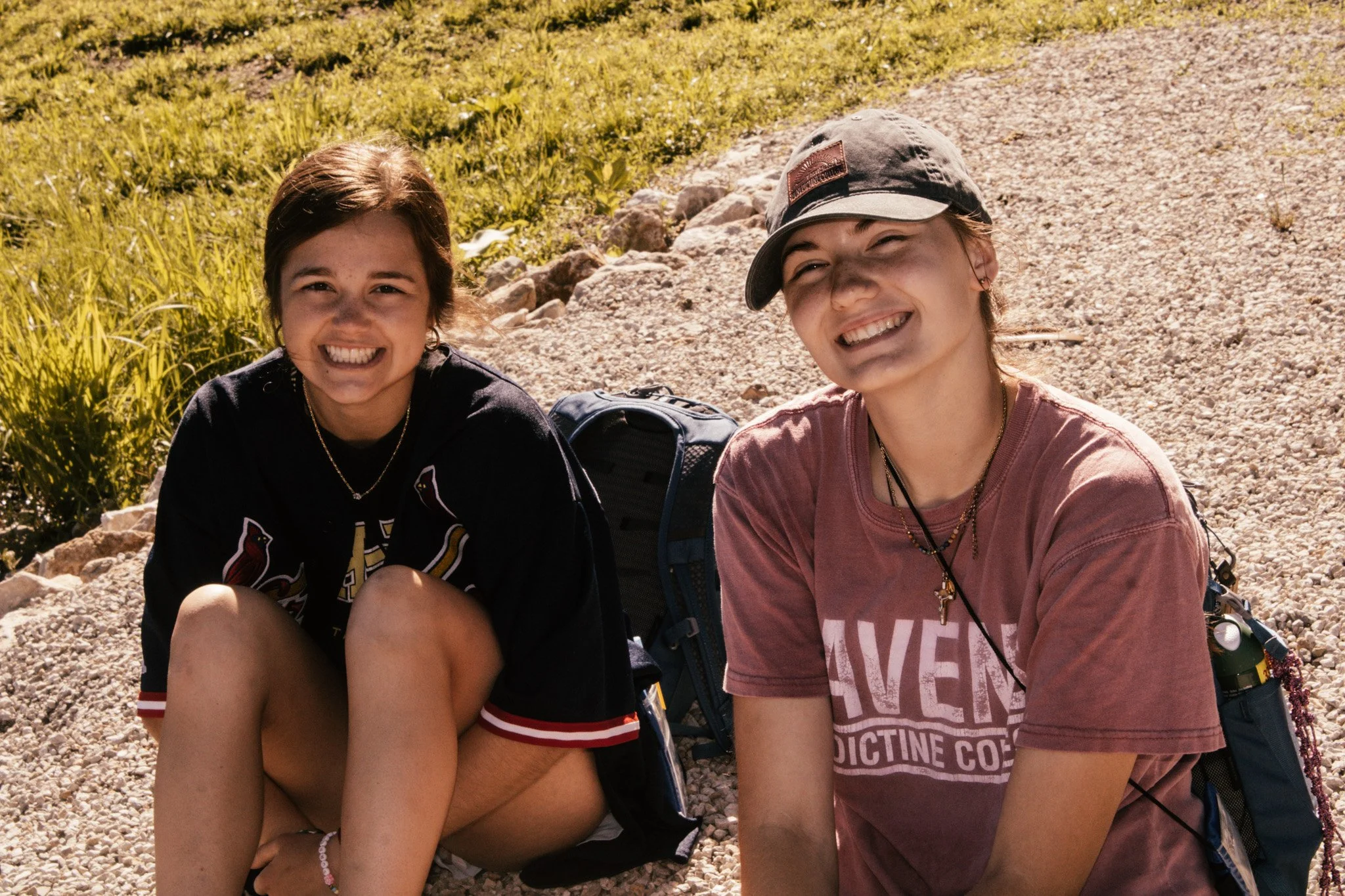 Two young girls smiling, sitting outdoors on rocky ground, with green grass and bushes in the background.
