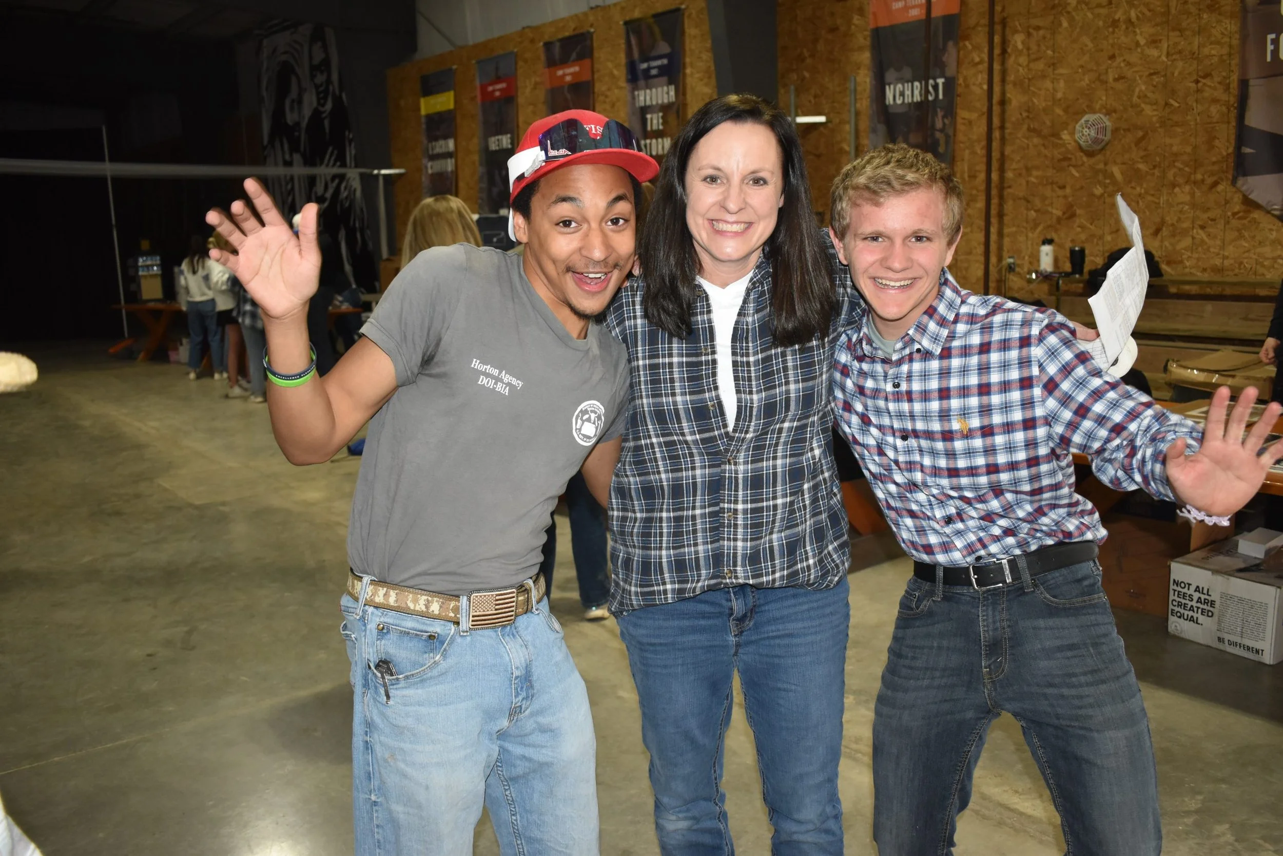 Three people smiling and posing with their hands raised inside a room with wooden walls and banners hanging in the background.