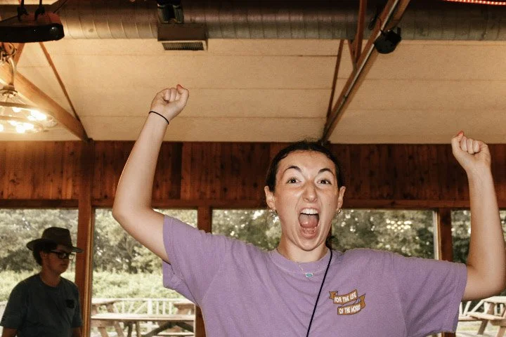 A woman in a purple shirt is excited, with her mouth open and fists raised, in a room with wooden walls and large windows showing trees outside.