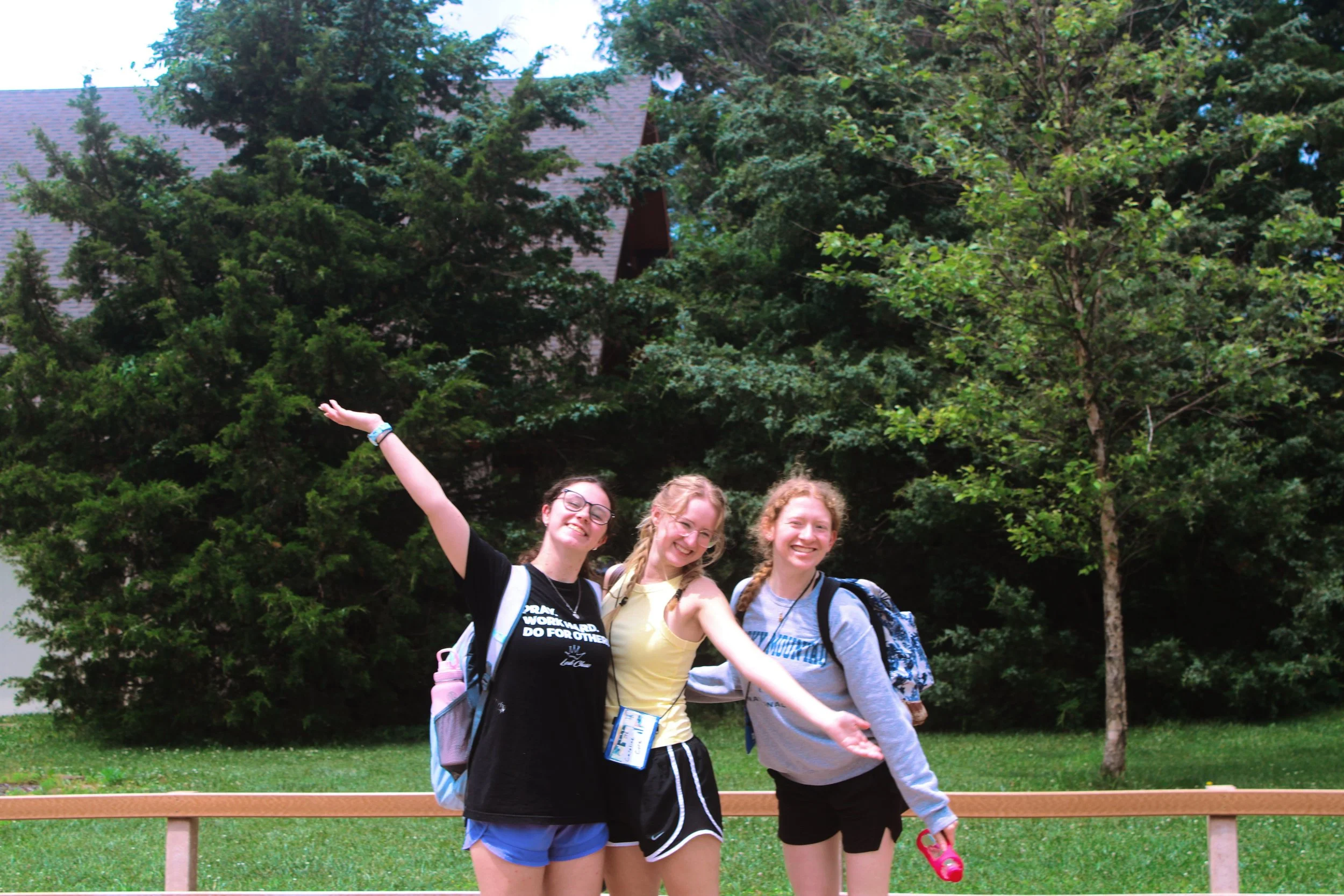 Four young women standing outdoors near a wooden railing, smiling and posing for a photo with trees and a house in the background.