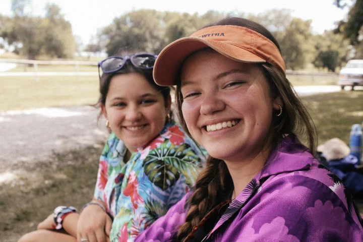Two young women sitting outside smiling at the camera, one wearing a colorful tropical shirt and sunglasses on her head, the other wearing a purple jacket and an orange cap.