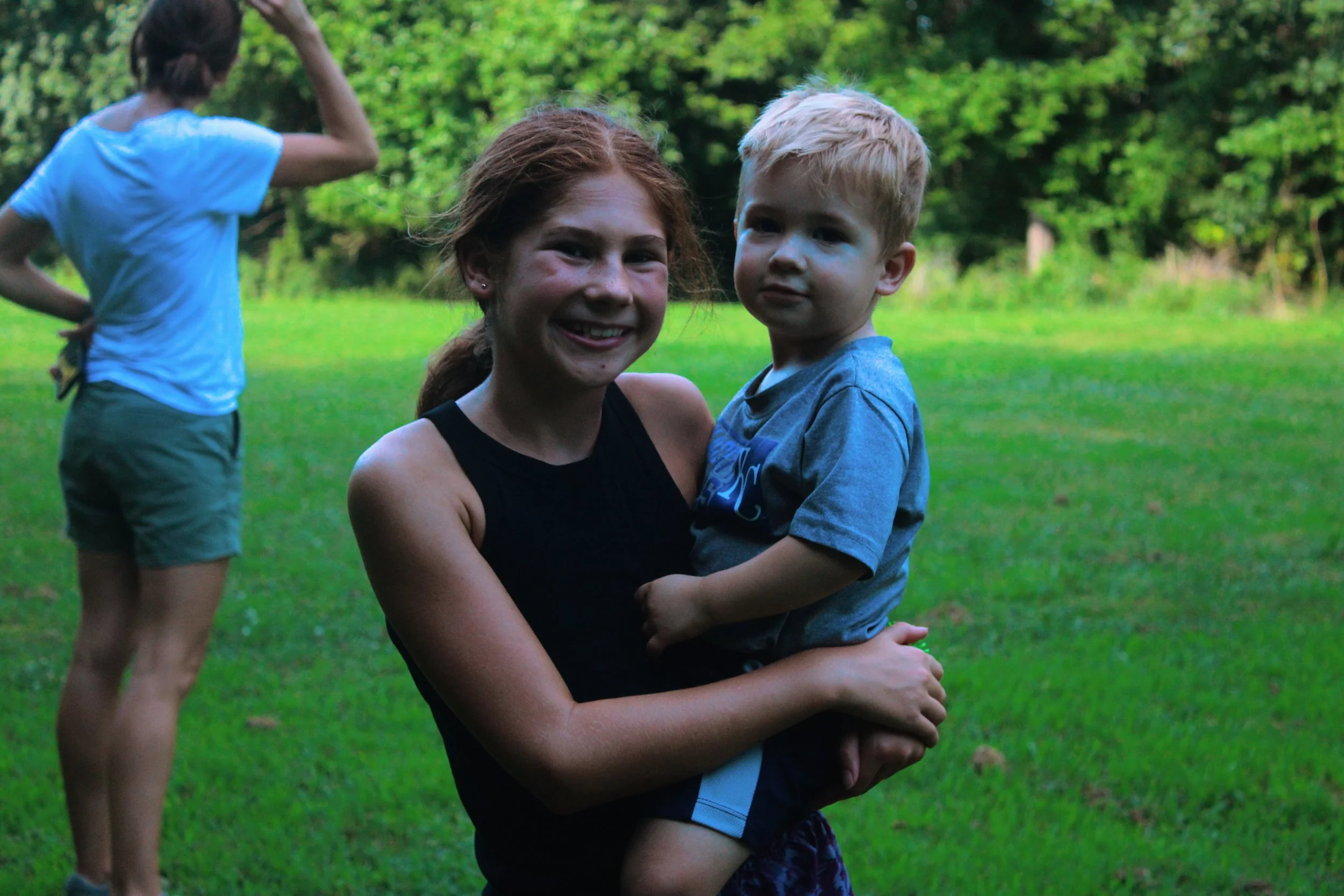 A woman with red hair smiling while holding a young blond boy in her arms outdoors in a park, with a green lawn and trees in the background. Another person with dark hair and a blue shirt is in the background, facing away.