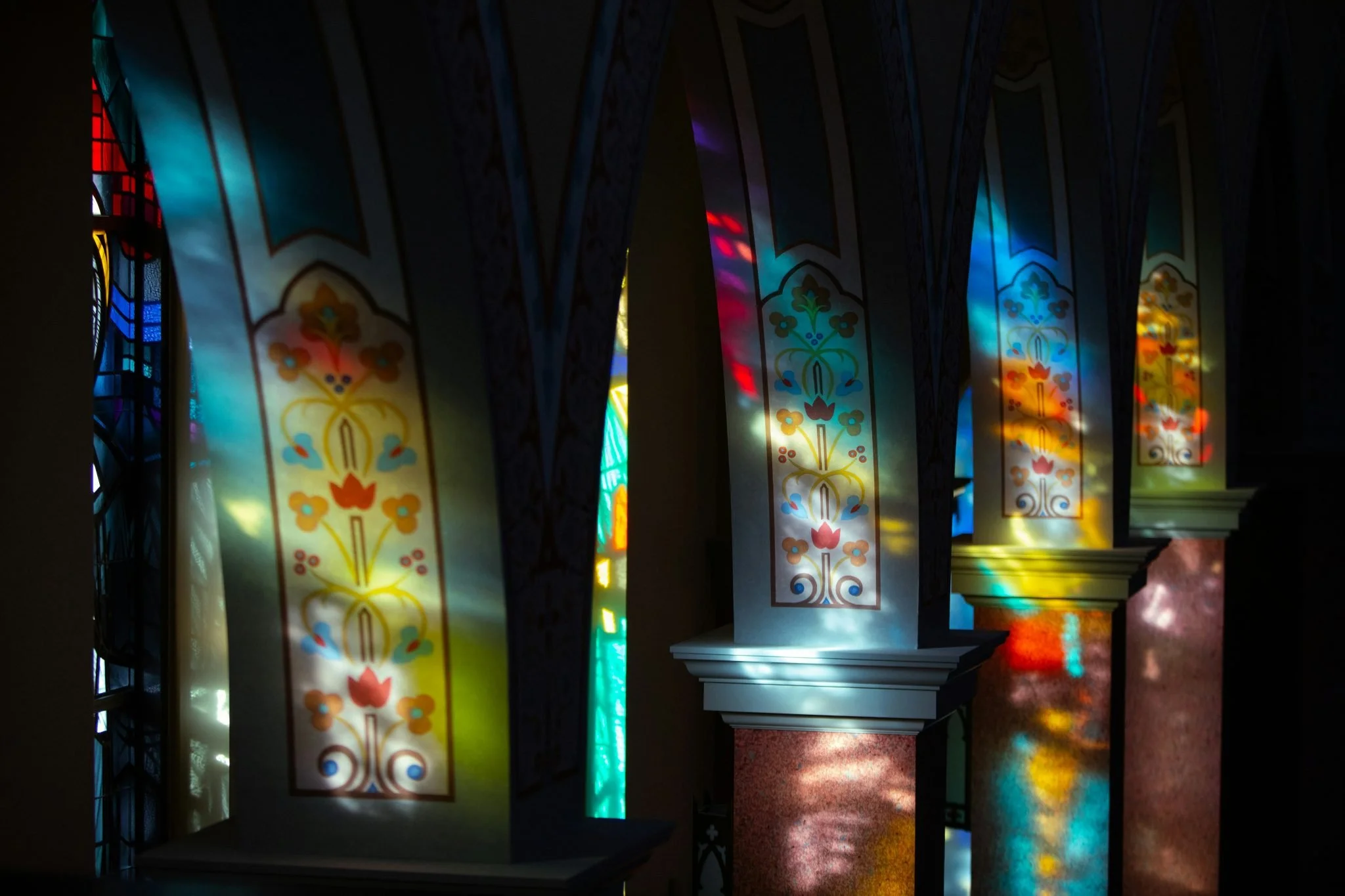 Colorful stained glass window panels with floral patterns illuminated by sunlight, creating vibrant reflections on the floor inside a building.