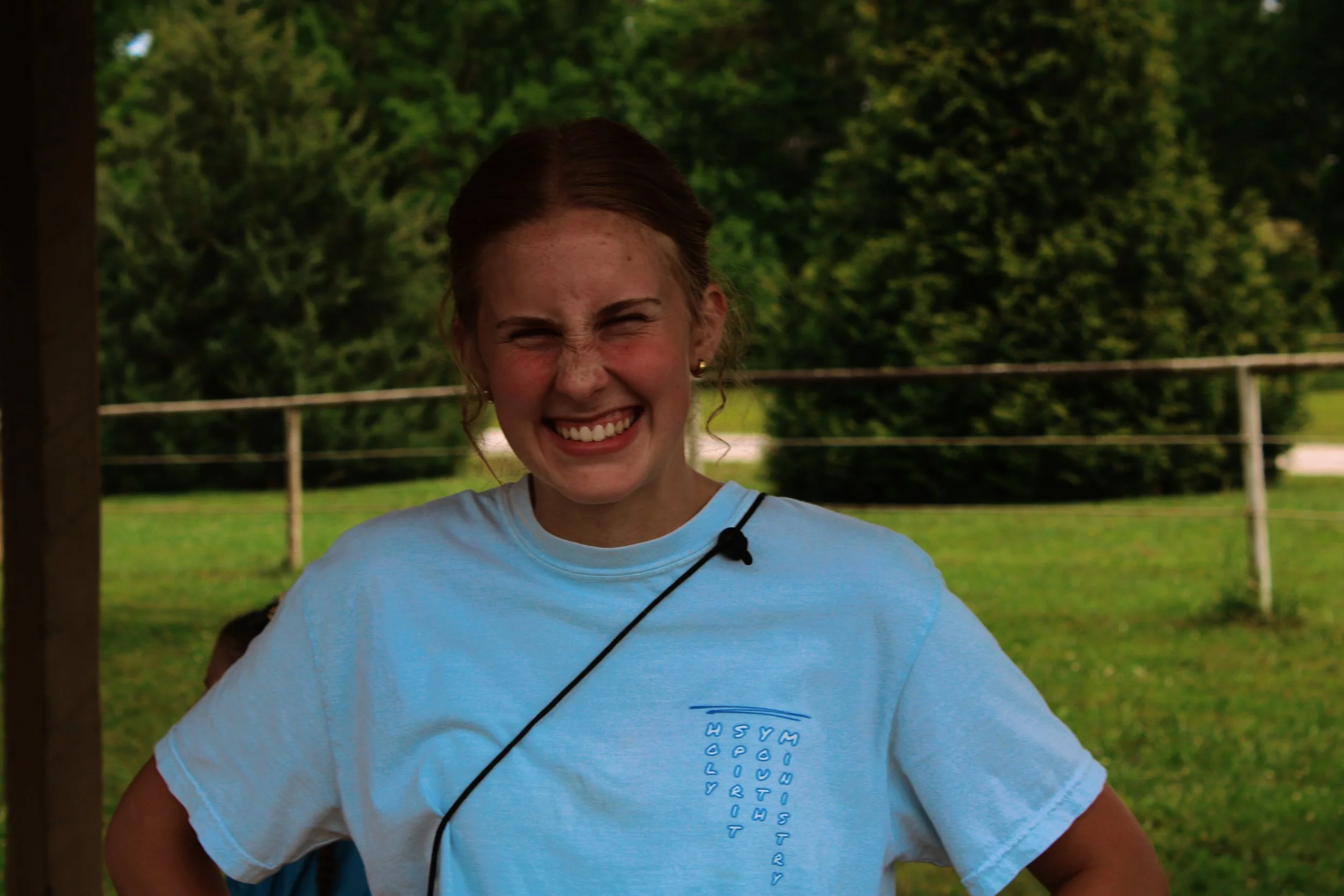 A young woman with red hair in a ponytail, smiling with her eyes closed, wearing a light blue shirt with a vertical line of text on it, standing outdoors with green trees and a wooden fence in the background.