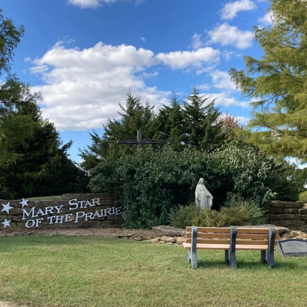 Memorial site with a sign that reads 'Mary Star of the Prairie', a statue of Mary, and a bench on a grassy area with trees and a partly cloudy sky in the background.