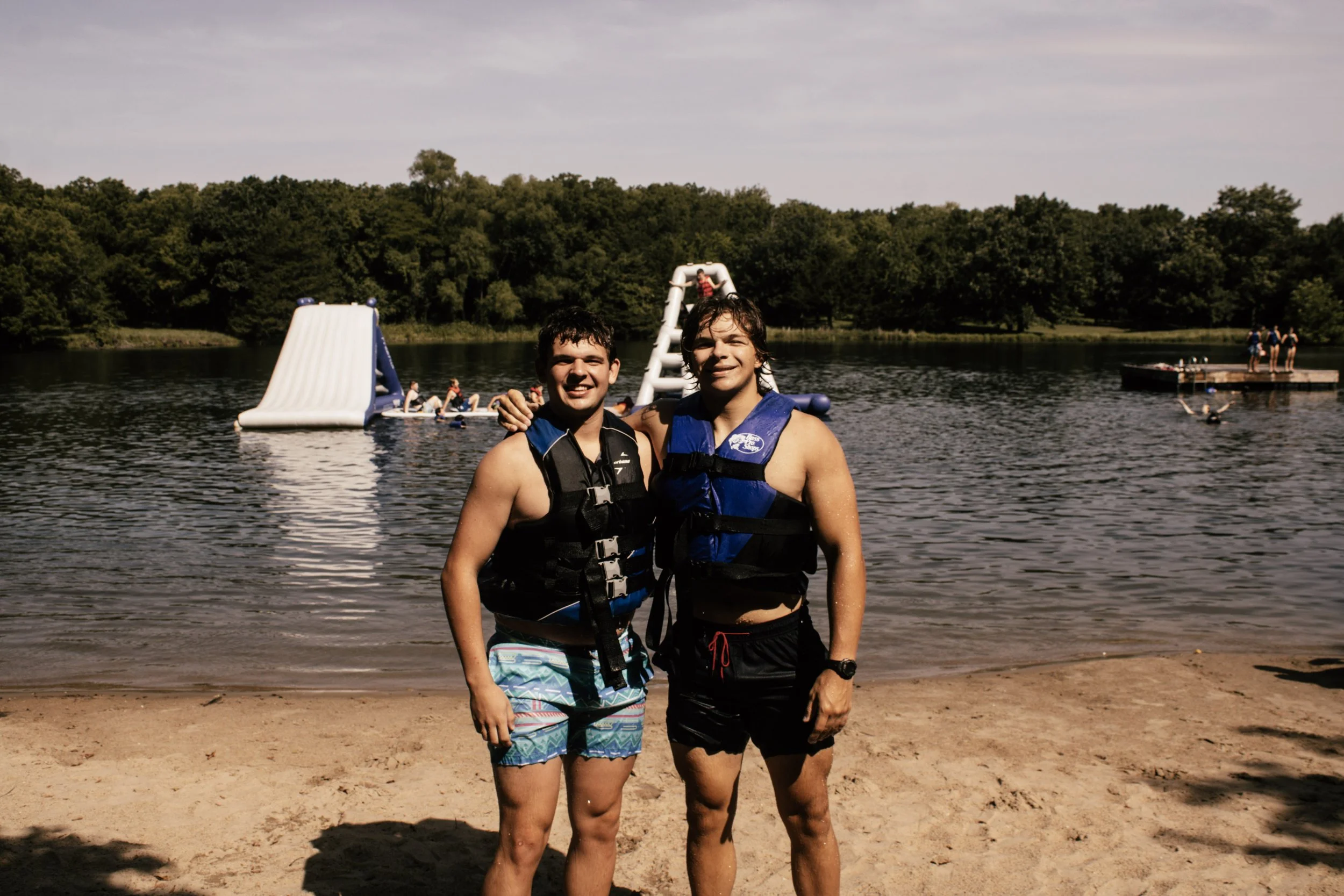 Two young men standing at the edge of a lake, smiling, wearing life jackets and swim trunks, with a dam and trees in the background and people swimming and playing in the water.