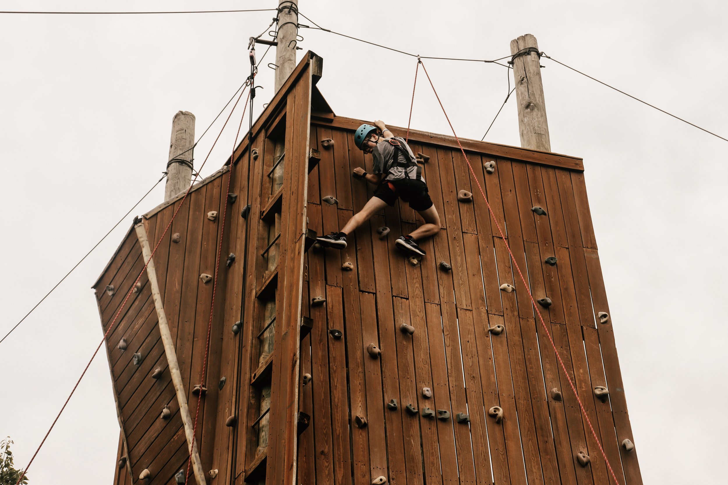 Young person in helmet climbing a wooden rock climbing wall outdoors.