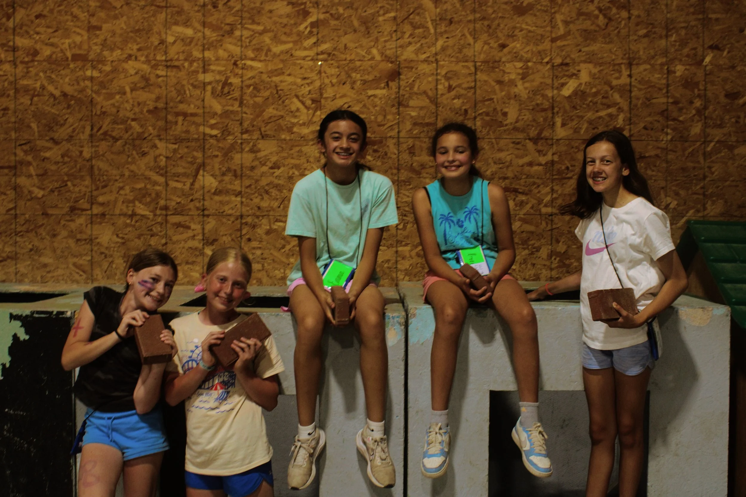 Group of six smiling young girls sitting on a ledge against a wooden wall, holding bricks, with some wearing bracelets and wearing casual athletic clothing.