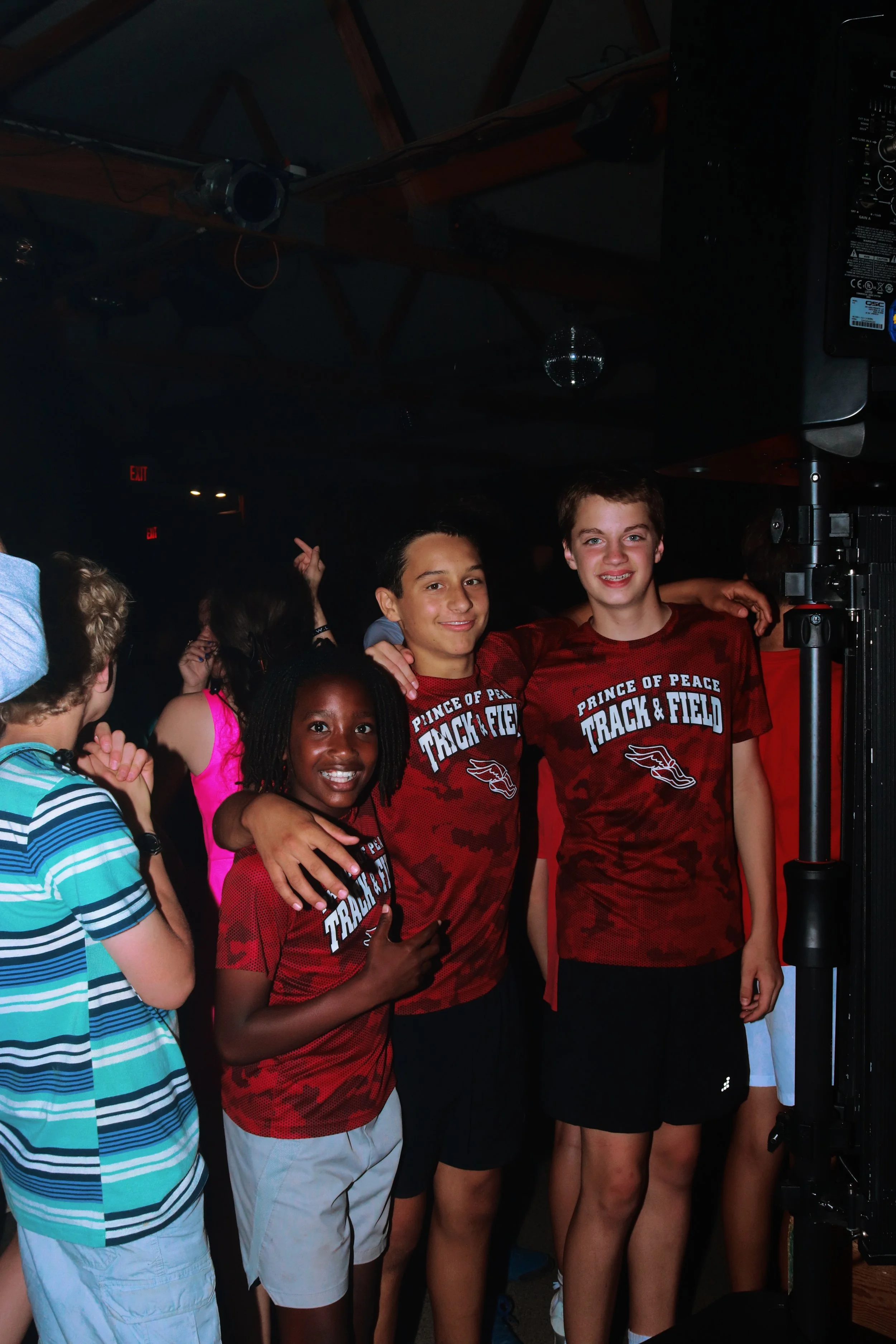 Group of teenagers at an indoor event, smiling and posing for a photo, wearing red sports shirts with 'Prince of Peace Track & Field' logo.