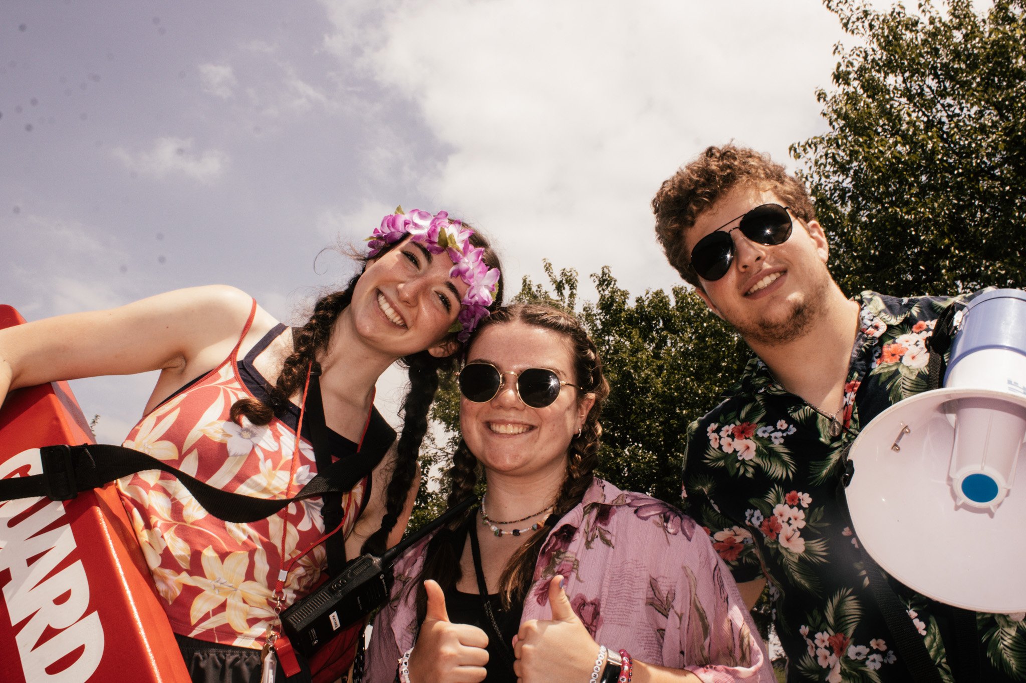 Four people smiling and posing for a photo outdoors. One woman wearing a flower crown and graphic dress, a woman with glasses and braids, a man with dark sunglasses and floral shirt, and a woman giving thumbs up, with trees and cloudy sky in the back