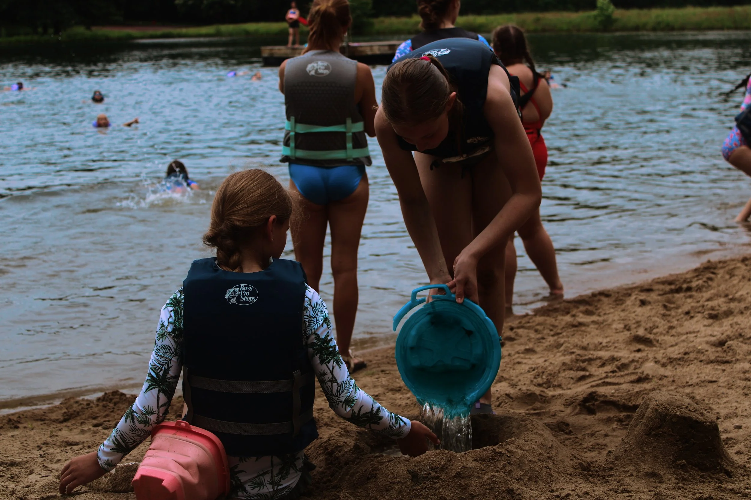 Children and adults playing on a sandy beach by a lake, with some kids swimming in the water and others filling buckets with sand, during daytime.