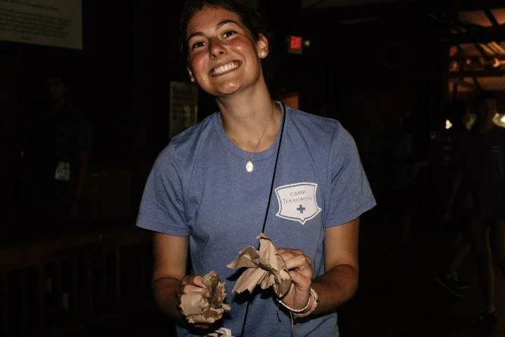 A young woman smiling while holding crumpled paper, wearing a blue camp T-shirt with a patch that says 'Camp TewaWk Wi' and a cross symbol, in a dimly lit indoor setting.