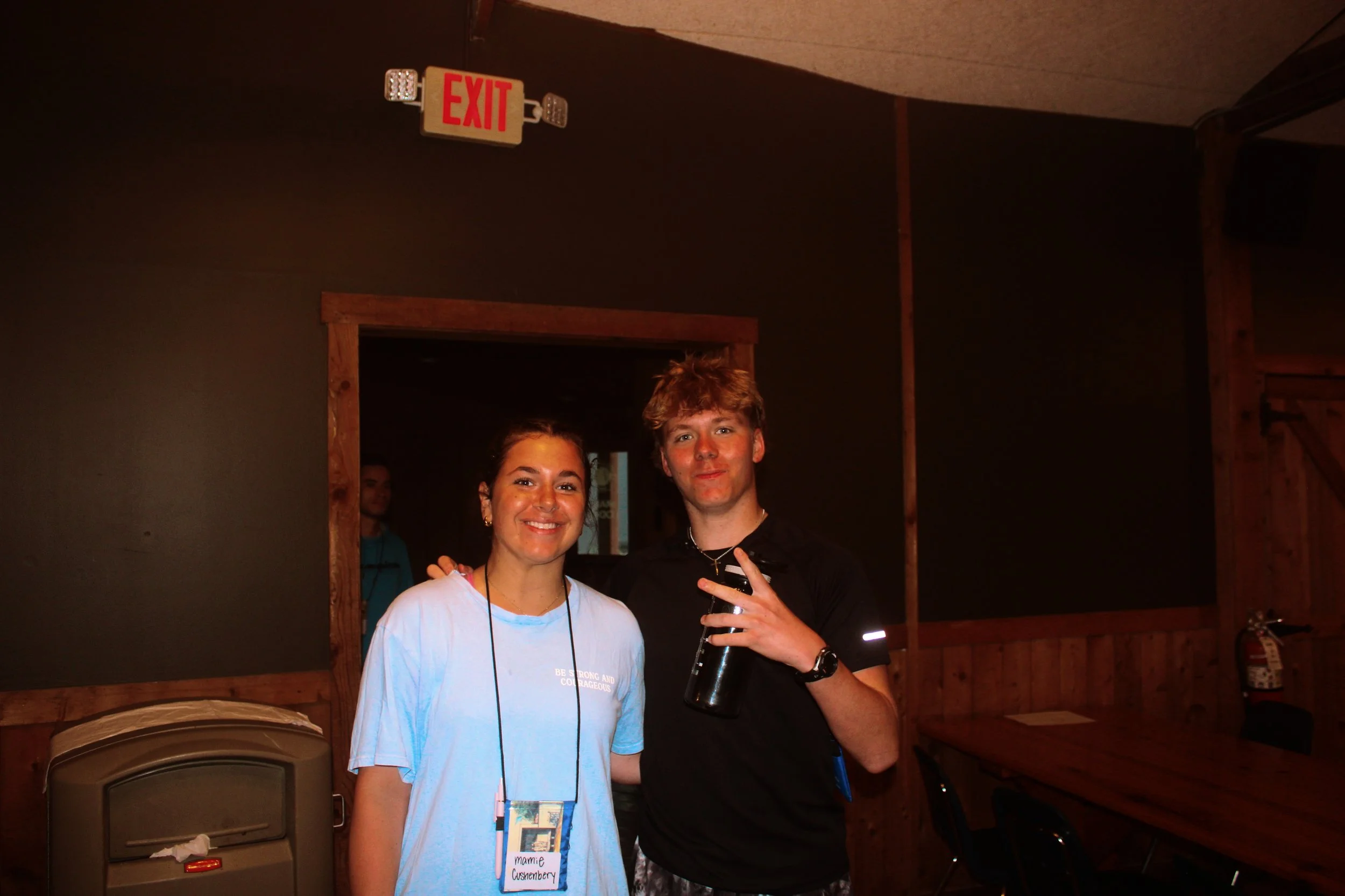 A woman and a man standing side by side inside a room with wooden walls. The woman is smiling and wearing a light blue shirt with a name tag that reads "Marnie Cushenberry." The man is holding a bottle of wine and making a peace sign. An exit sign an