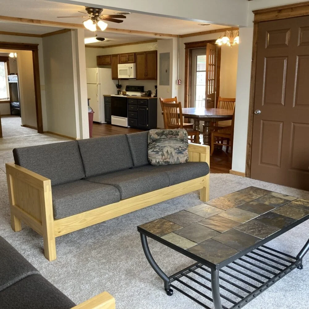 Living room with a grey sofa, a coffee table with a tile top, and a ceiling fan with lights. In the background, there is a dining area with a round wooden table and chairs, and a kitchen with white and black cabinets, white appliances, and wooden trim.