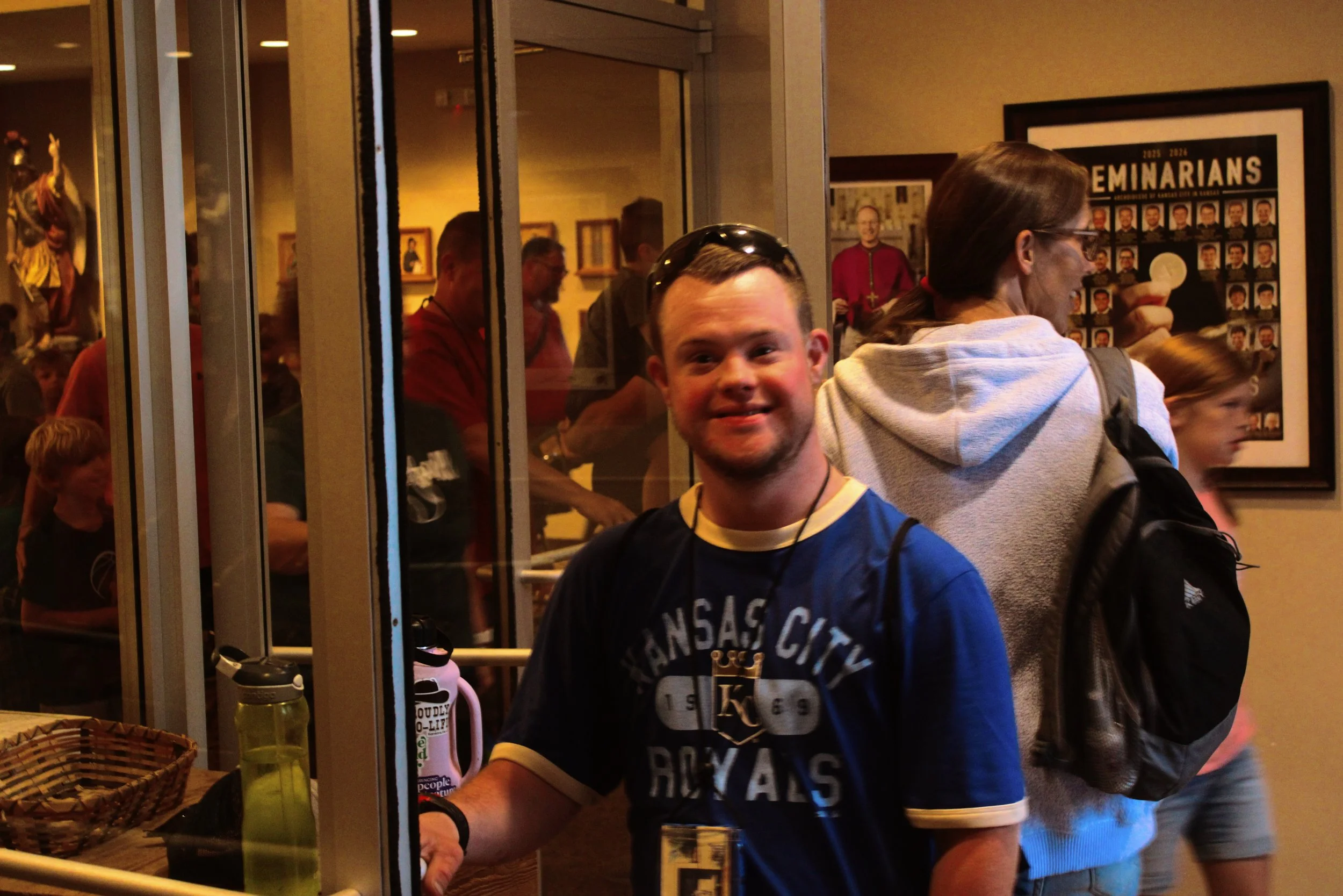 People gathered behind a glass window, some are standing, with framed pictures on the wall and a colorful statue visible in the background.
