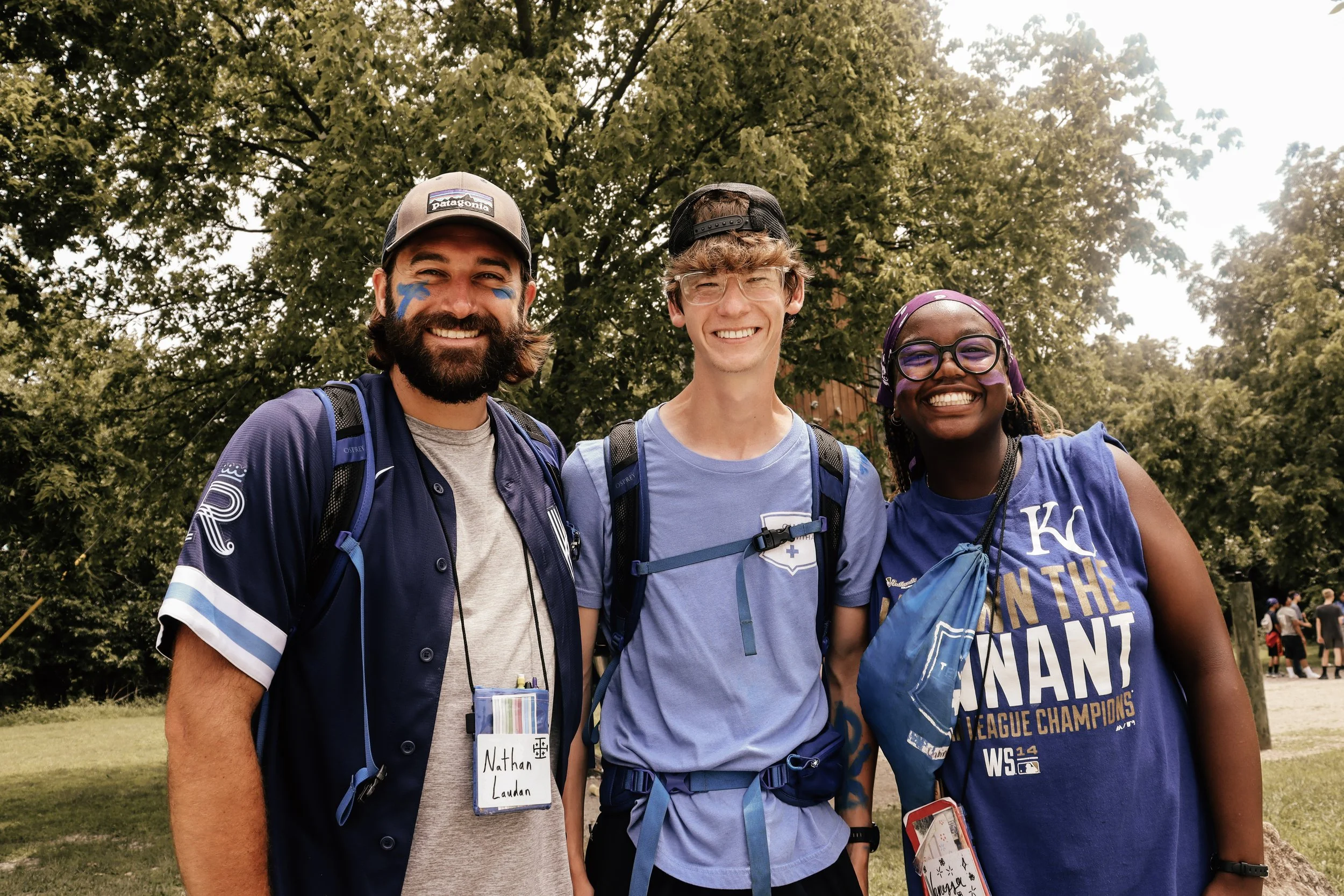 Three smiling people standing outdoors among trees, with two men on the left and a woman on the right, all wearing athletic clothing and carrying backpacks.