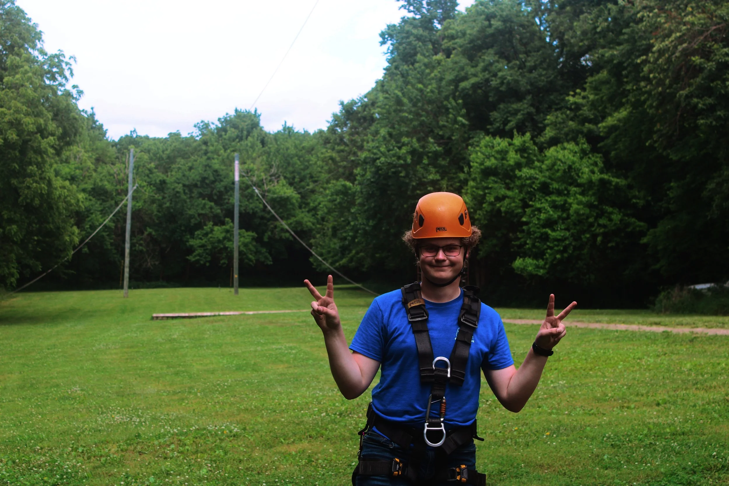 Person wearing an orange climbing helmet and a harness standing on a grassy field, smiling and making peace signs with both hands, with trees and power lines in the background.