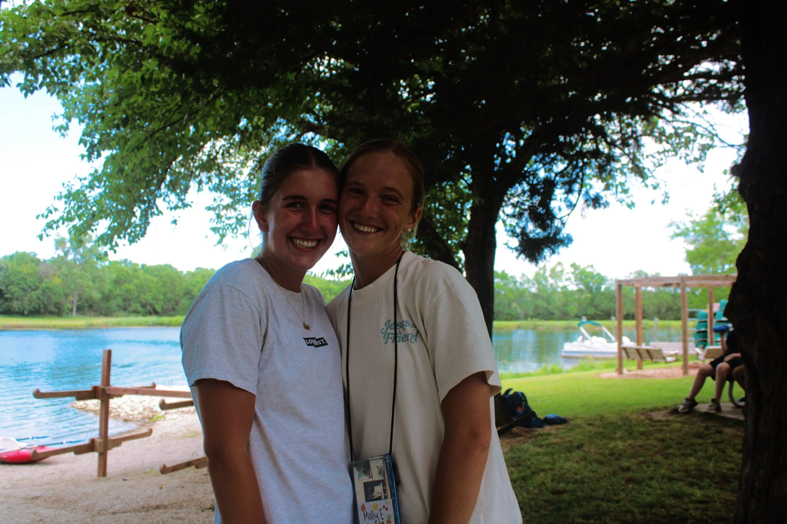 Two smiling young women standing closely together outdoors near a lake, with trees and a wooden dock in the background on a sunny day.