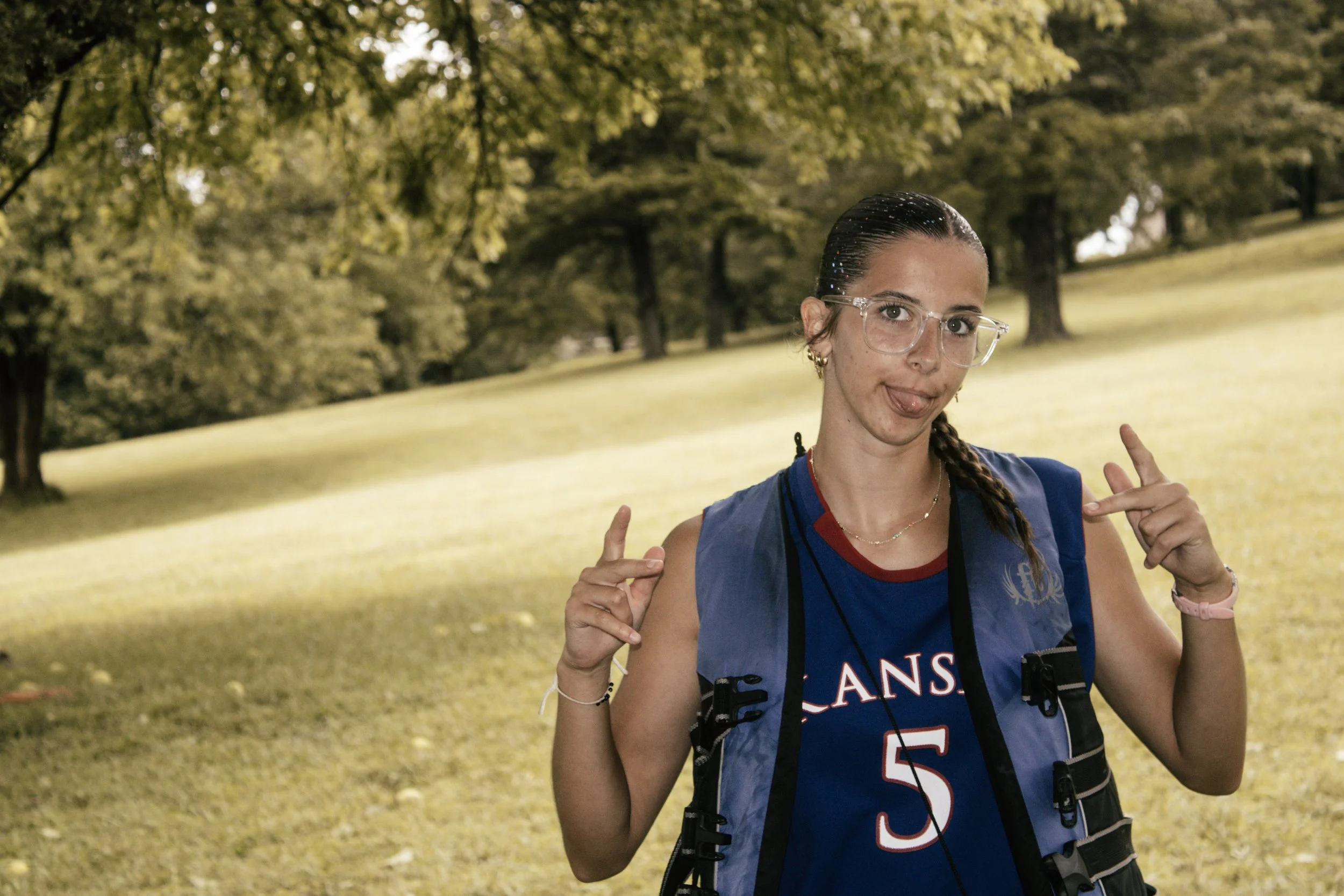 A woman in athletic gear, wearing glasses and a sports jersey with 'LANS' visible, making a peace sign with her fingers in a park with trees and grass.