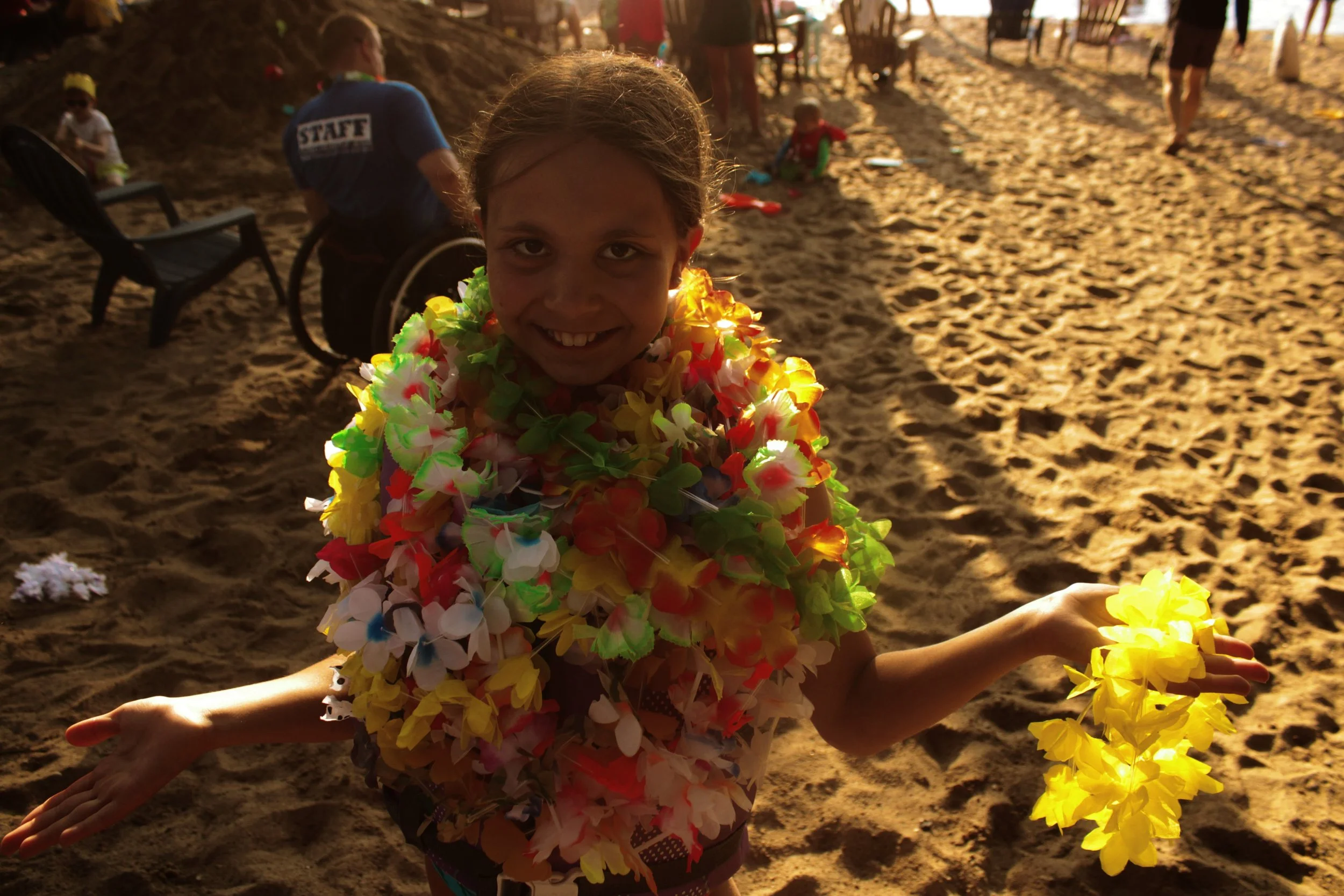 A young girl smiling at the camera, wearing a colorful lei made of flower leis, with her arms extended, at a beach or sandy area during sunset. In the background, people are sitting and playing on the sand, some with leis, with warm golden light illu