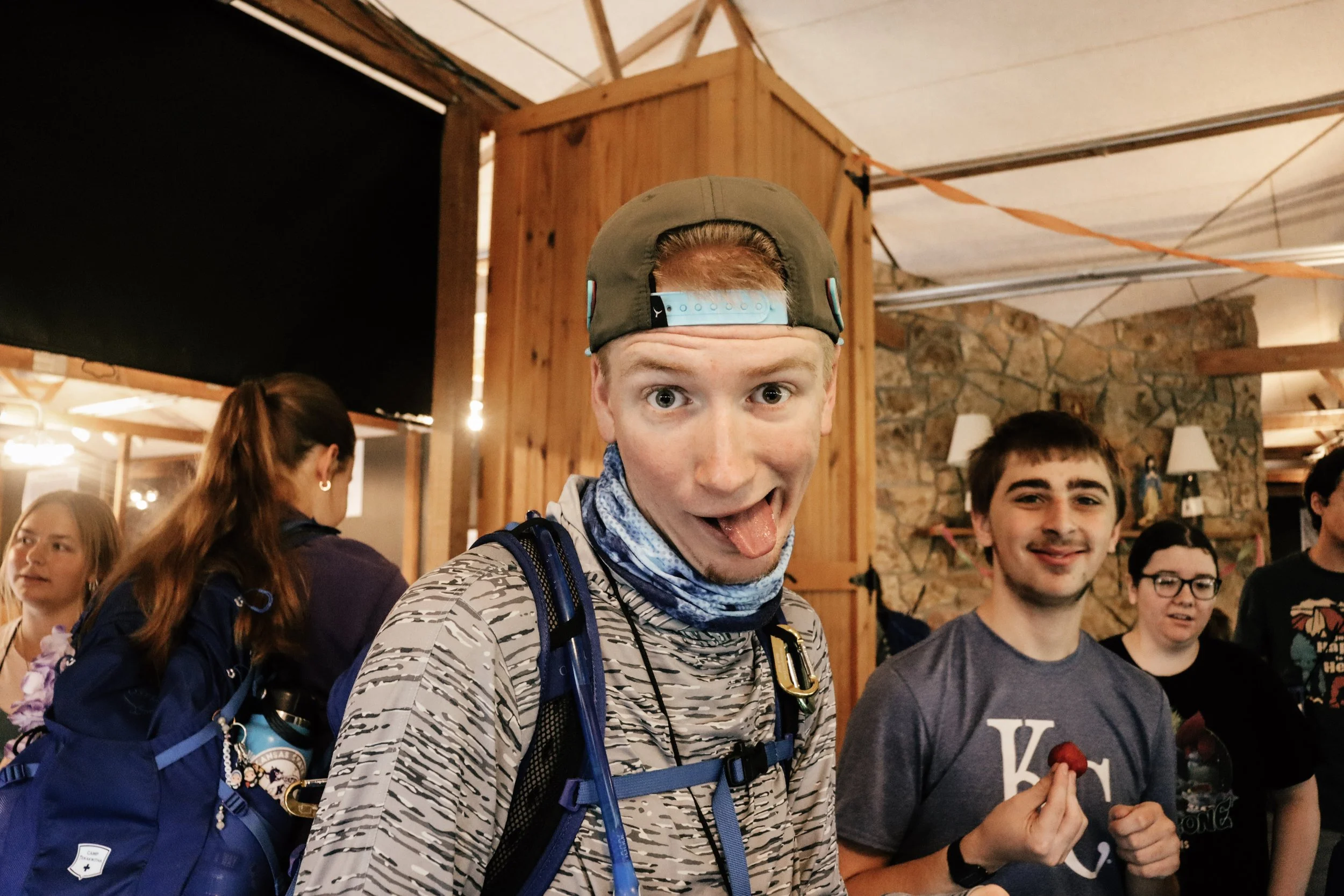 A young man with red hair wearing a baseball cap backwards, making a funny face with his tongue out. He has a backpack and scarf around his neck, and is surrounded by other smiling young people indoors with wooden walls and brick decor.
