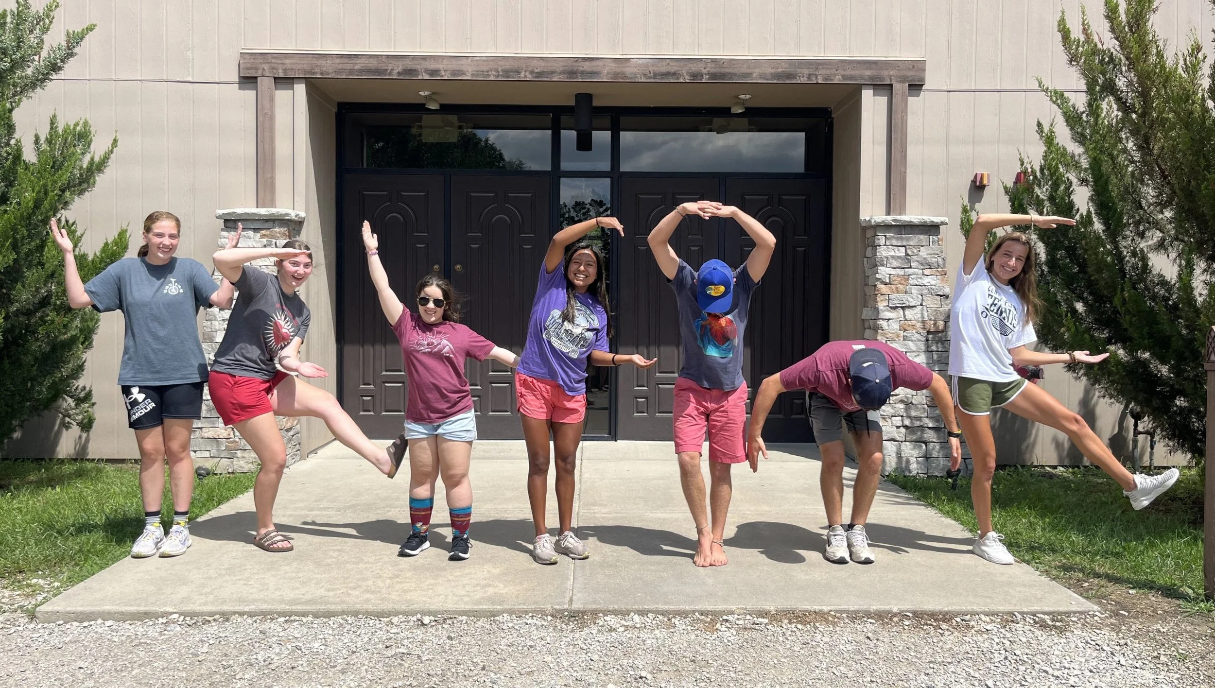 Group of young people posing playfully in front of a building, some making yoga poses with trees on either side.