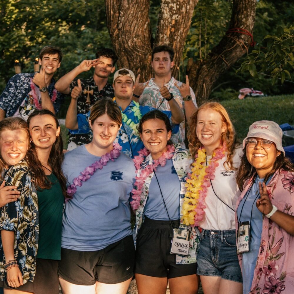 Group of teenagers and women smiling outdoors near a tree, wearing leis and casual summer clothes, some with name tags, during daytime.