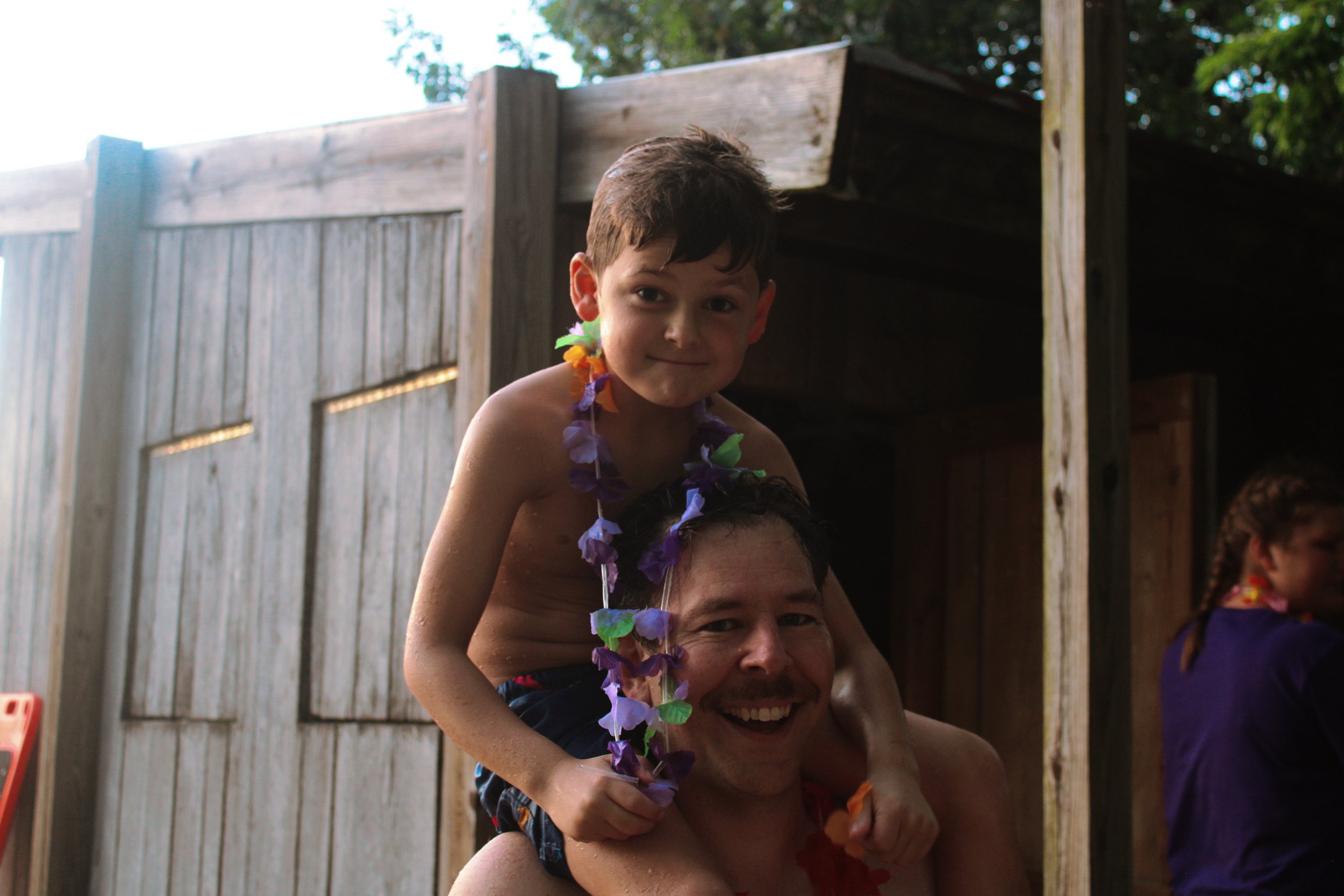 A young boy sitting on a man's shoulders during a backyard celebration, wearing a colorful lei, with wooden fencing and trees in the background.