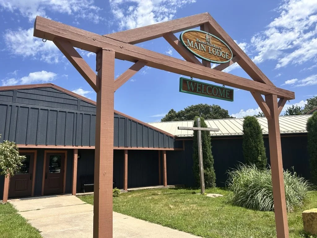 Sign for Main Lodge with a welcome sign underneath, with a building in the background, trees, and a partly cloudy sky.