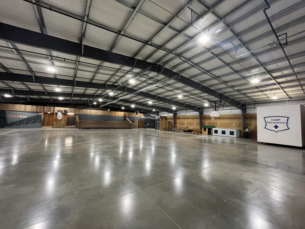 Empty indoor gymnasium with polished concrete floor, high ceiling, basketball hoop, and various wooden structures and signs, including one labeled 'Camp Tekawitha' and another labeled 'black box'.