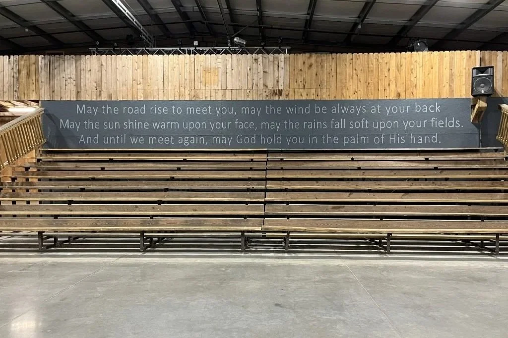 Empty wooden bleacher seats in a gymnasium or auditorium with a sign displaying a prayer on the wall behind.