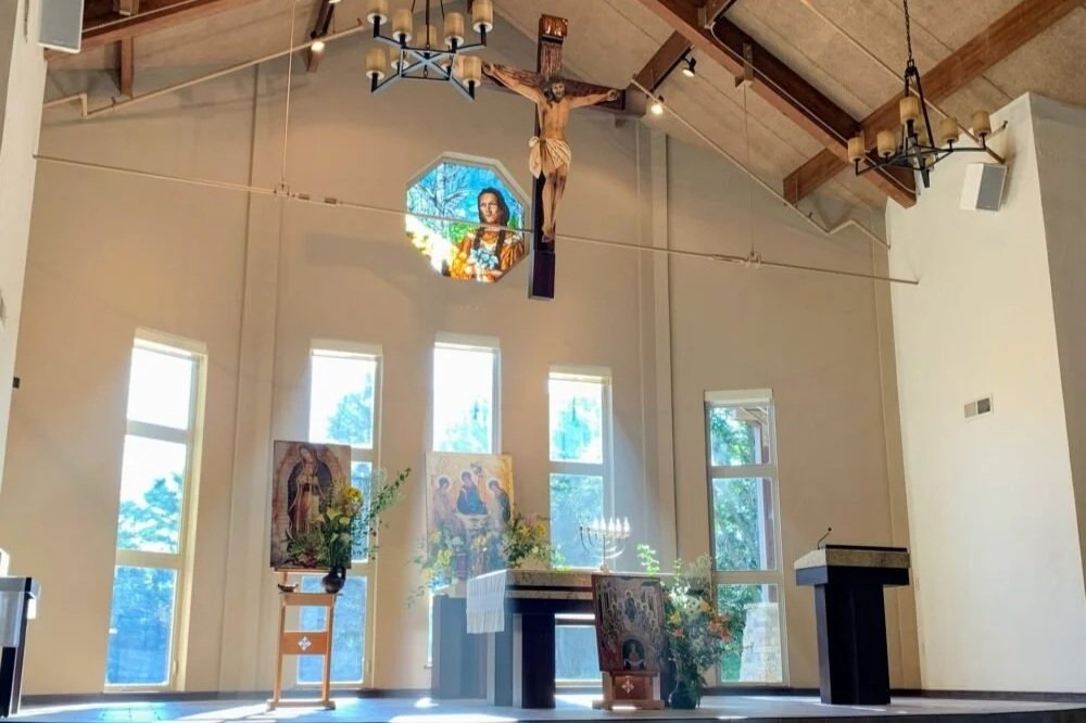 Interior of a church altar with religious icons, a crucifix hanging from the ceiling, and stained glass windows.