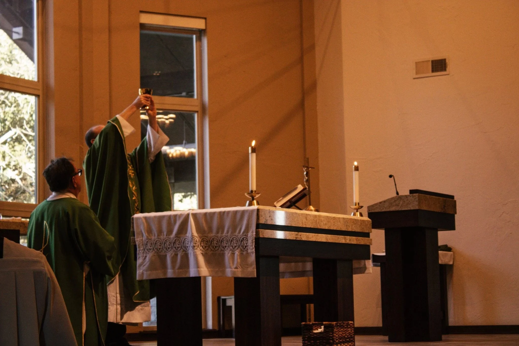 A person dressed in green liturgical robes holds up a chalice in a church during a ceremony, with other individuals also dressed in similar robes nearby.