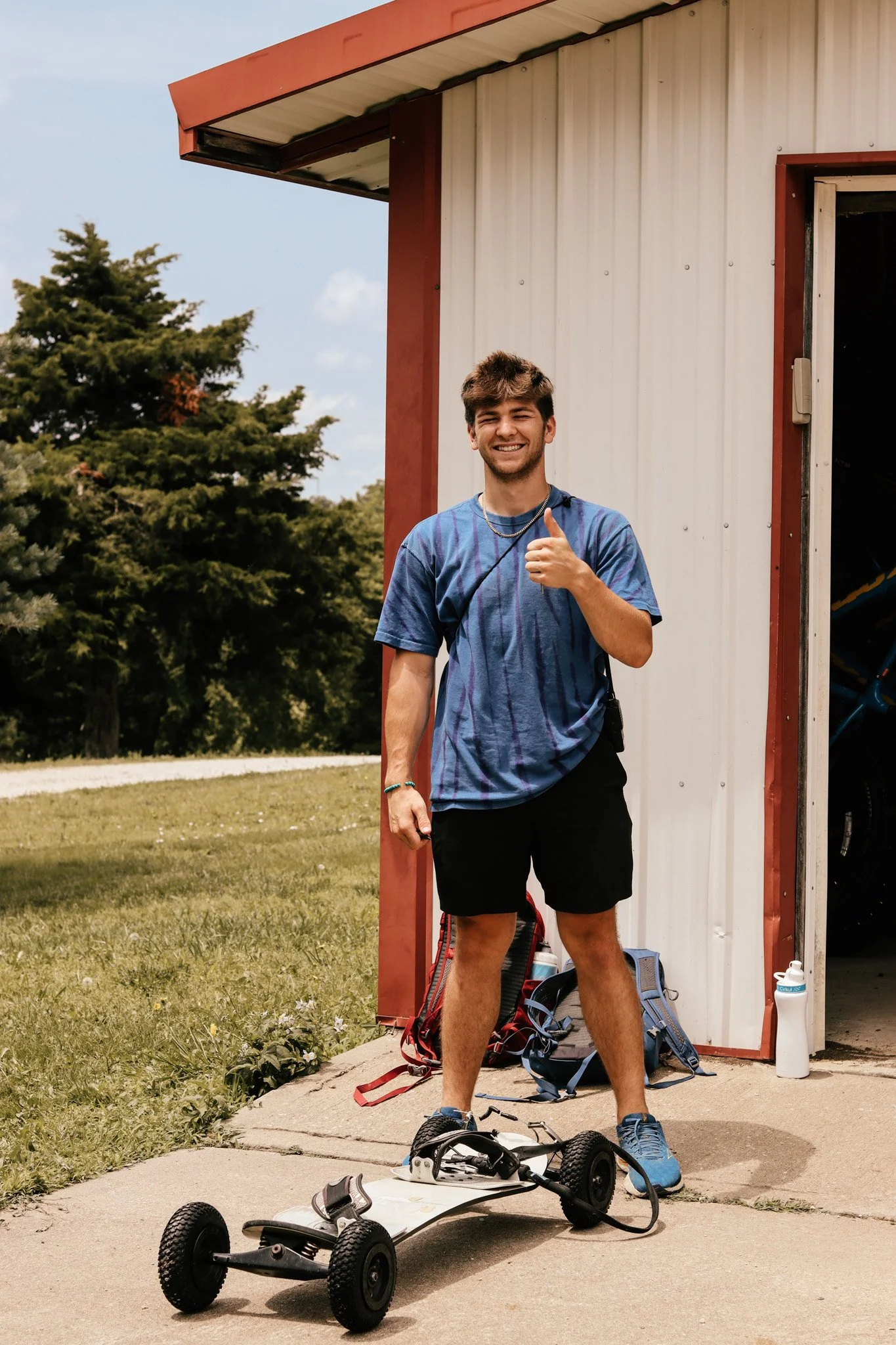 A young man with a big smile, wearing a blue t-shirt, black shorts, and blue sneakers, stands next to an electric skateboard on a sidewalk outside a building with a red border. He is giving a thumbs-up gesture. There are backpacks, a water bottle, an