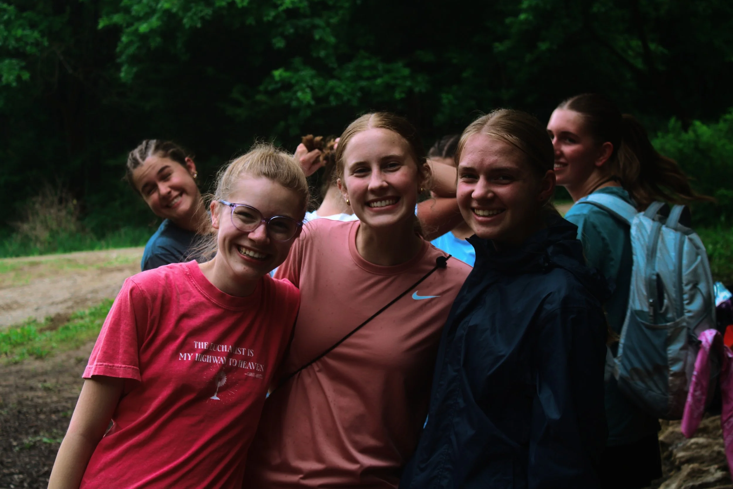 A group of six smiling young teenage girls in colorful casual clothing, standing close together outdoors in a wooded area, enjoying a day in nature.