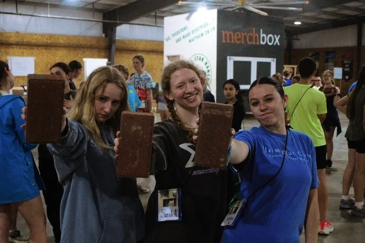 Three young women standing in a busy indoor event holding large brown rectangular objects, smiling at the camera.
