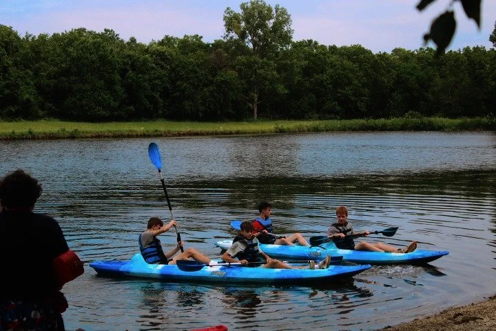 Four children kayaking on a calm river, with trees lining the shore in the background.