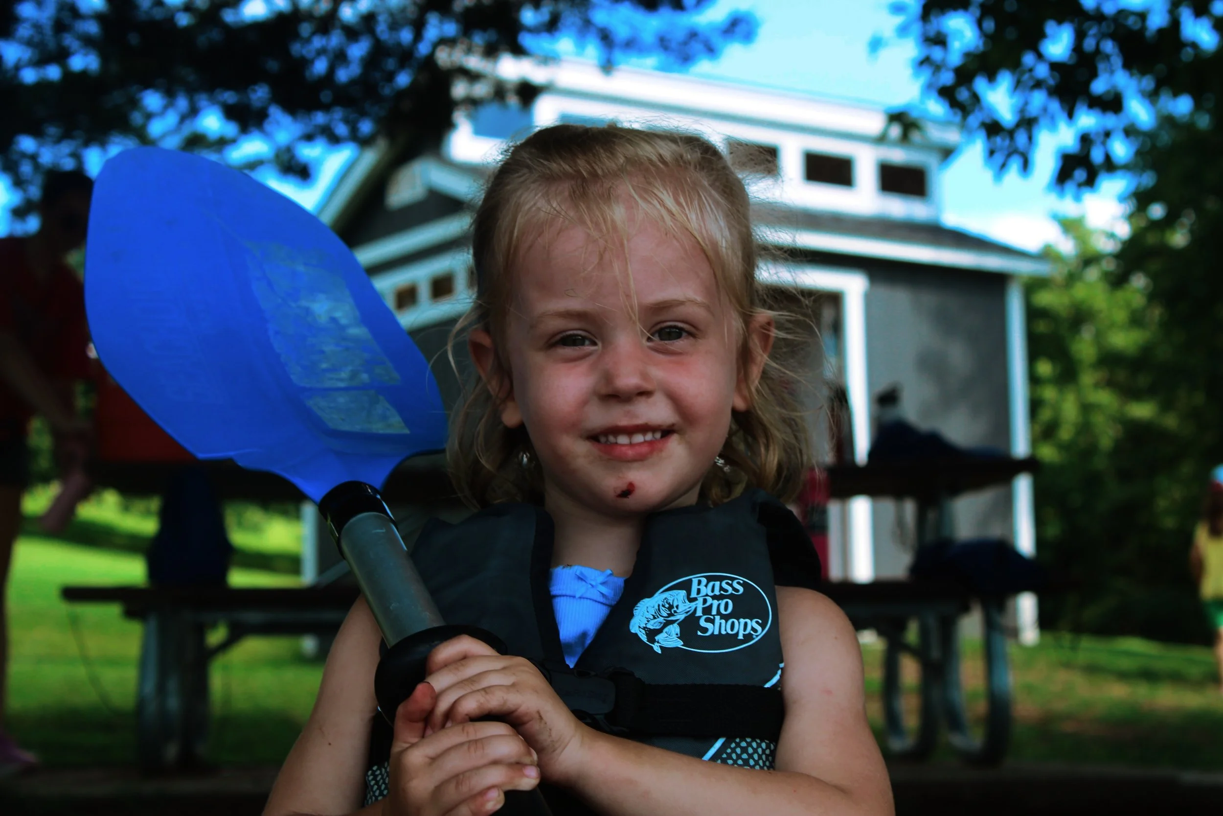 A young girl with blonde hair smiling while holding a blue fishing net outdoors, with green trees and a house in the background.