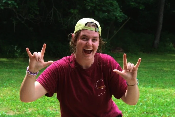 A young woman wearing a yellow backward cap and a red t-shirt smiling and making rock and roll hand gestures outdoors on green grass with trees in the background.