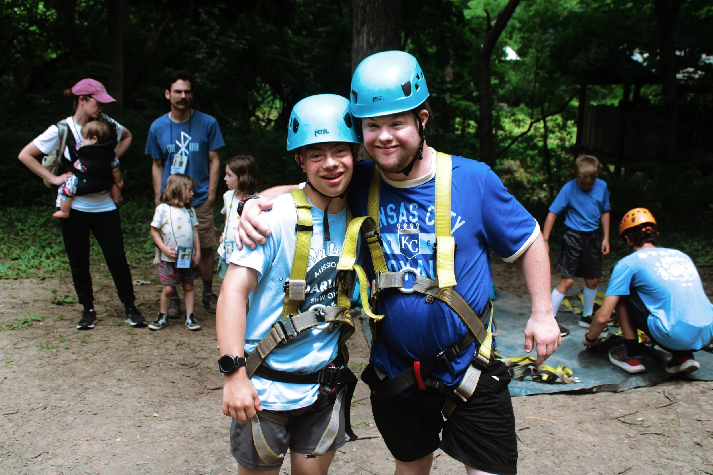 Two young men in blue shirts and helmets smiling with their arms around each other outdoors. In the background, a woman with a child in a carrier on her back, adults, and children are preparing for activity in a wooded area.