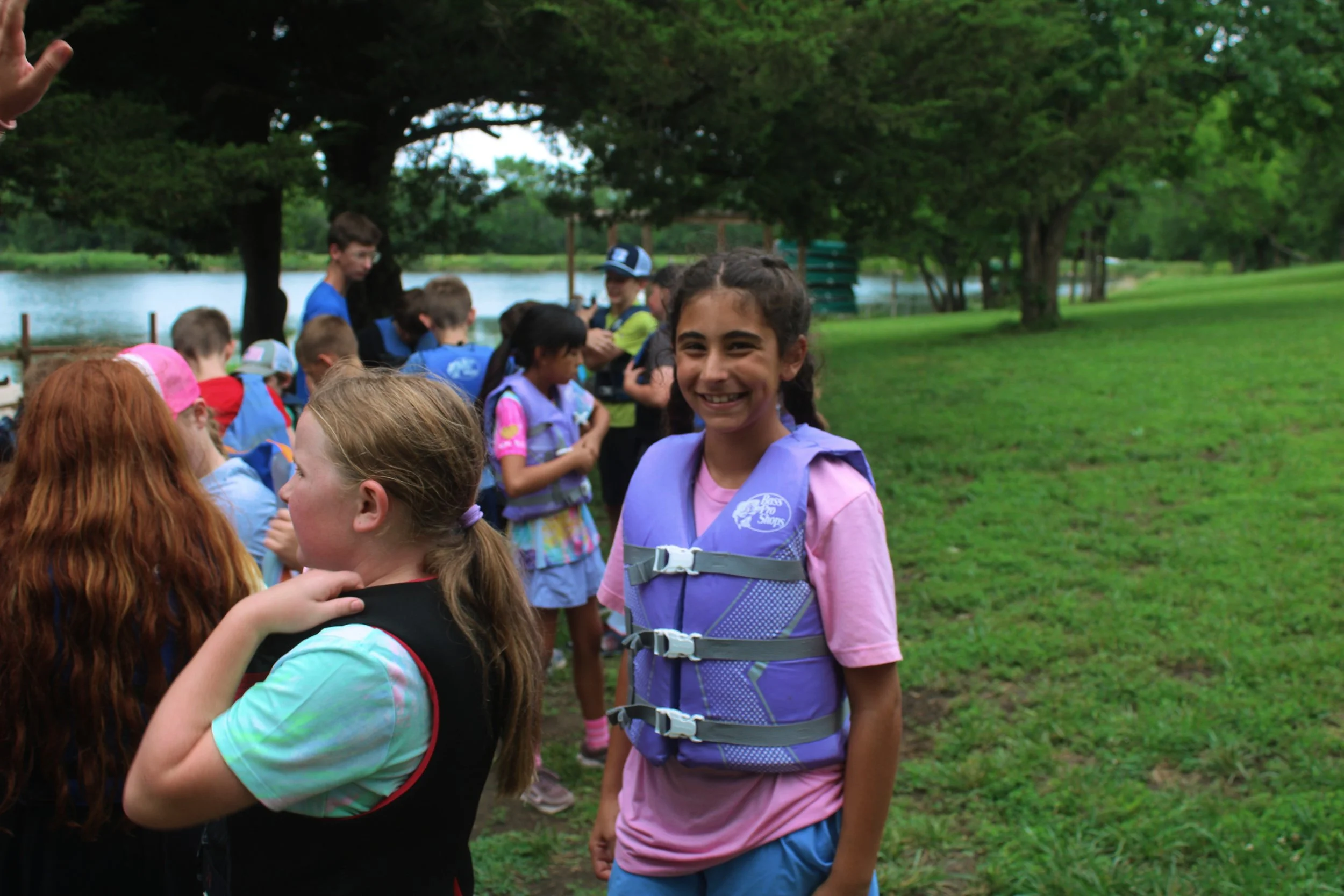 Smiling girl in pink t-shirt and purple life jacket standing outdoors near a lake, surrounded by children and adults.