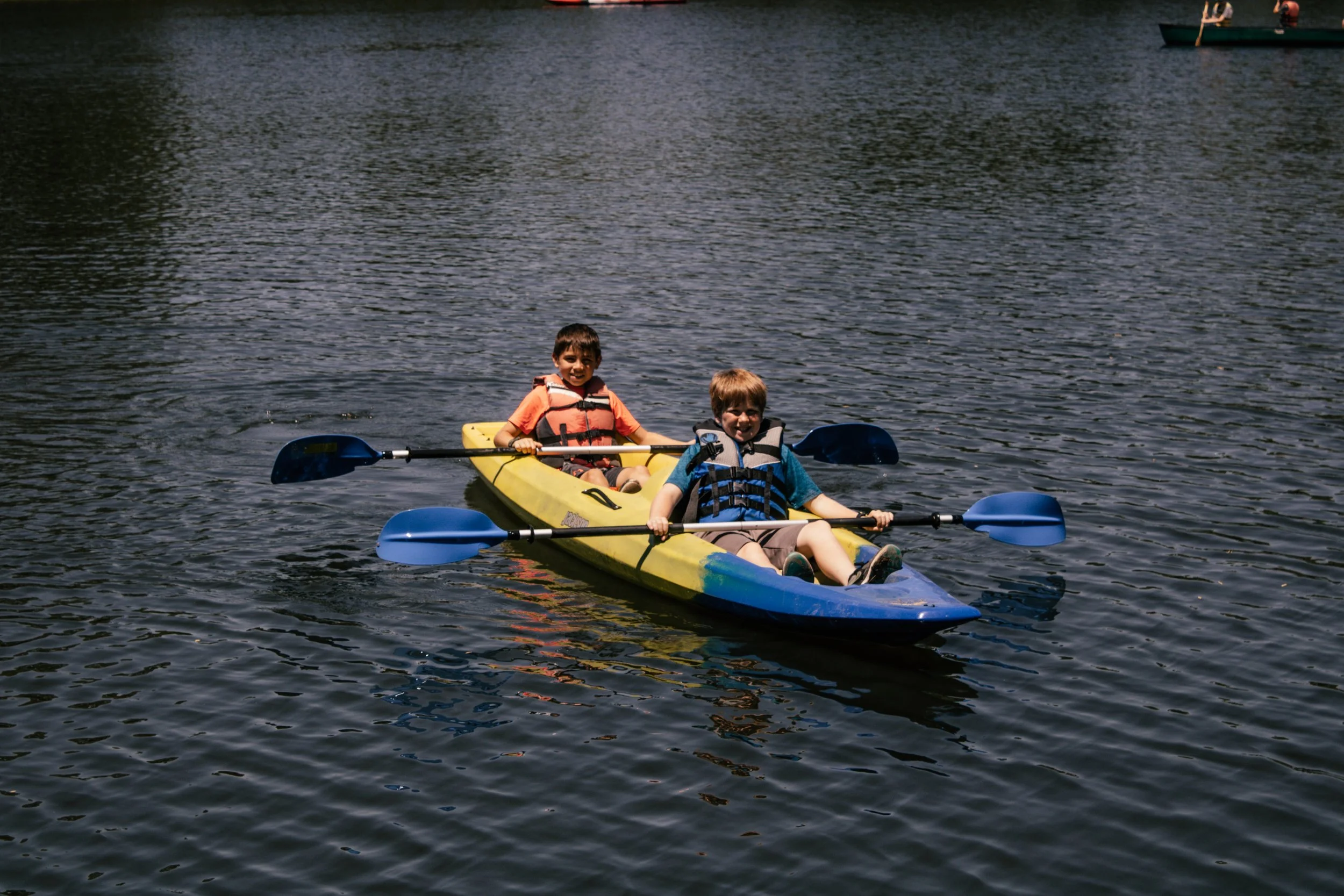 Two children paddling in a yellow kayak on a lake, wearing life jackets, and smiling.