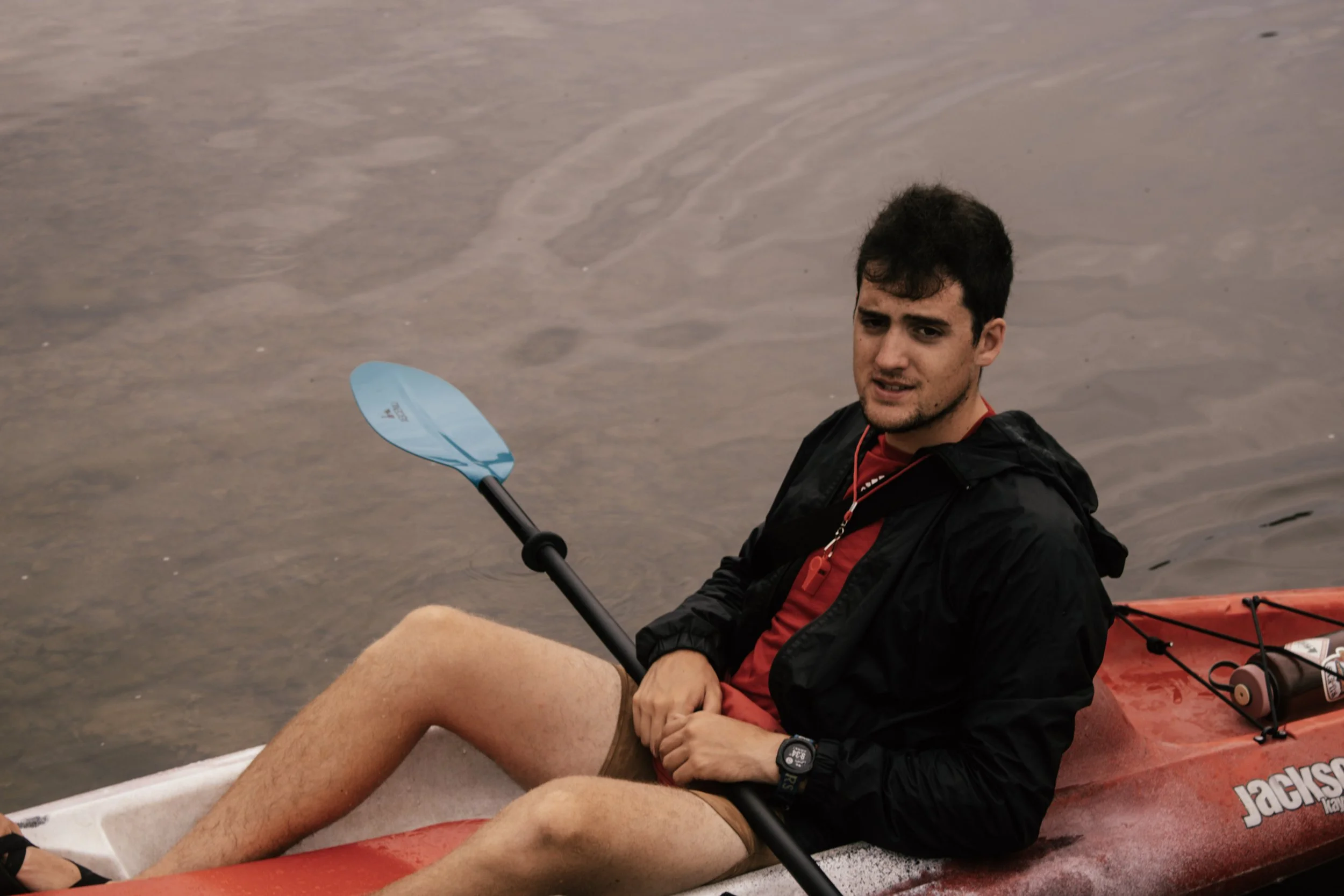 A young man with dark hair sitting in a kayak on a calm body of water, holding a paddle, wearing a black jacket and a red shirt underneath, with part of his leg visible in the foreground.
