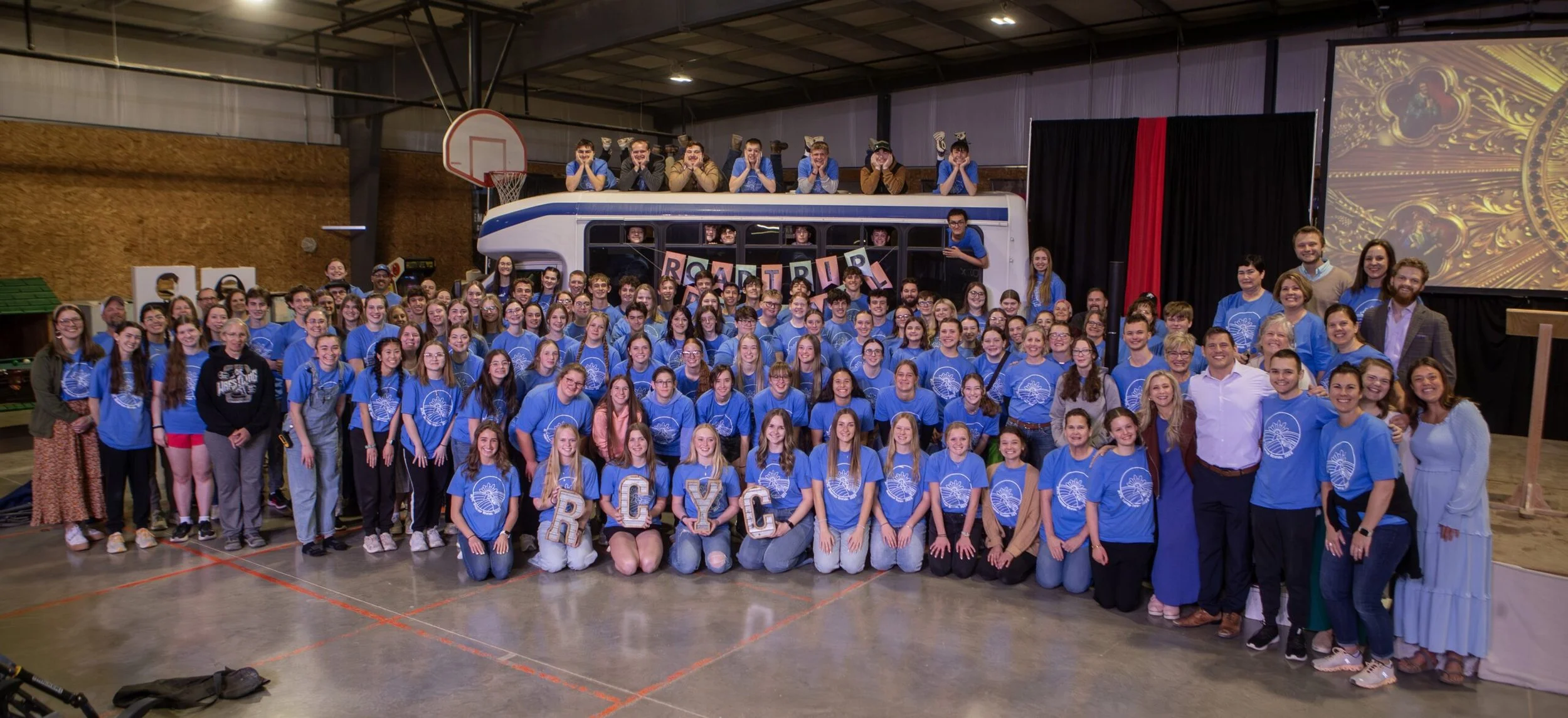 Group photo of a large crowd of young people and adults, many wearing matching blue t-shirts with a white emblem, inside a gymnasium with a basketball hoop and a stage. The stage has a bus prop with people leaning on top and a sign that spells 'RETRR